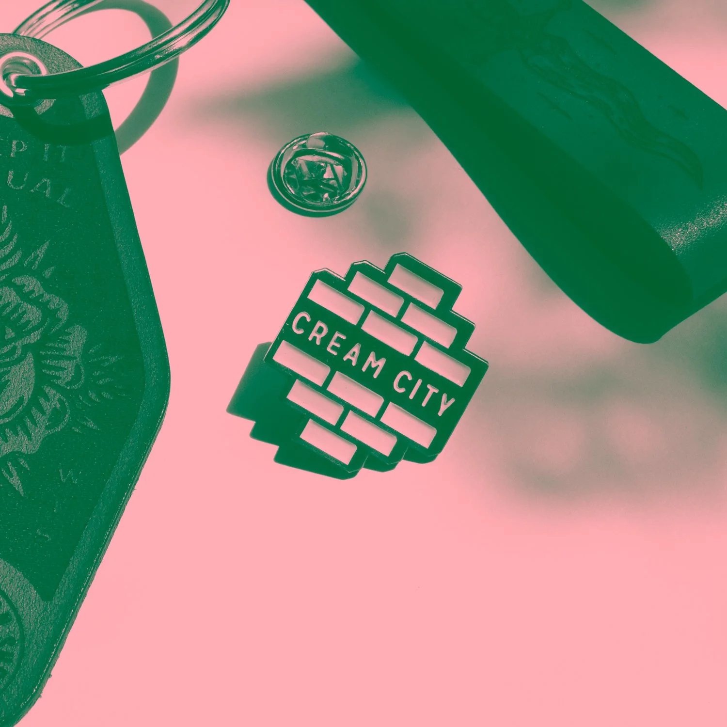 Close-up of a pink and black social media-style cutout sign reading 'CREAM CITY' with a brick wall pattern decorative background, a metal tin with a floral design, a green book or box, a black container, and a small metallic item on a pink surface.