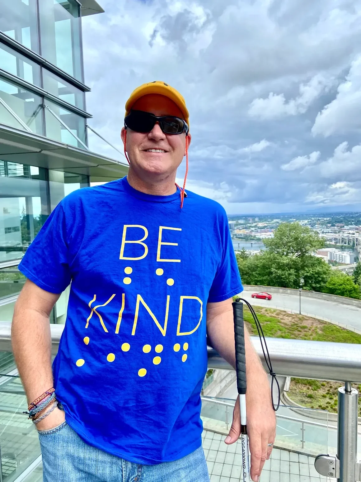 A white man wearing black sunglasses, a yellow hat, and a blue shirt that says "BE KIND" in the written alphabet and in braile, smiles at the camera and holds his cane