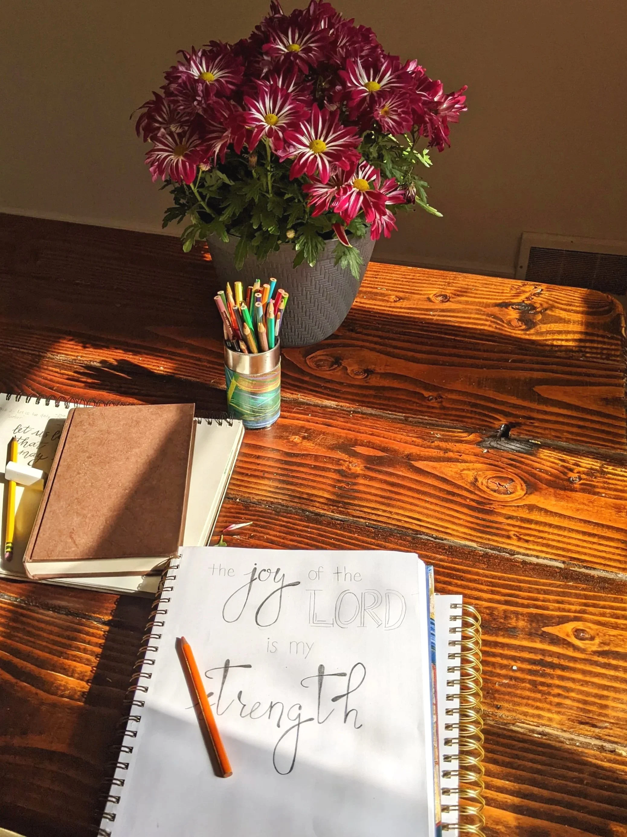 A wooden table with a vase of red and white flowers, a notebook, a journal, a pen, and a spiral note pad with handwritten text.