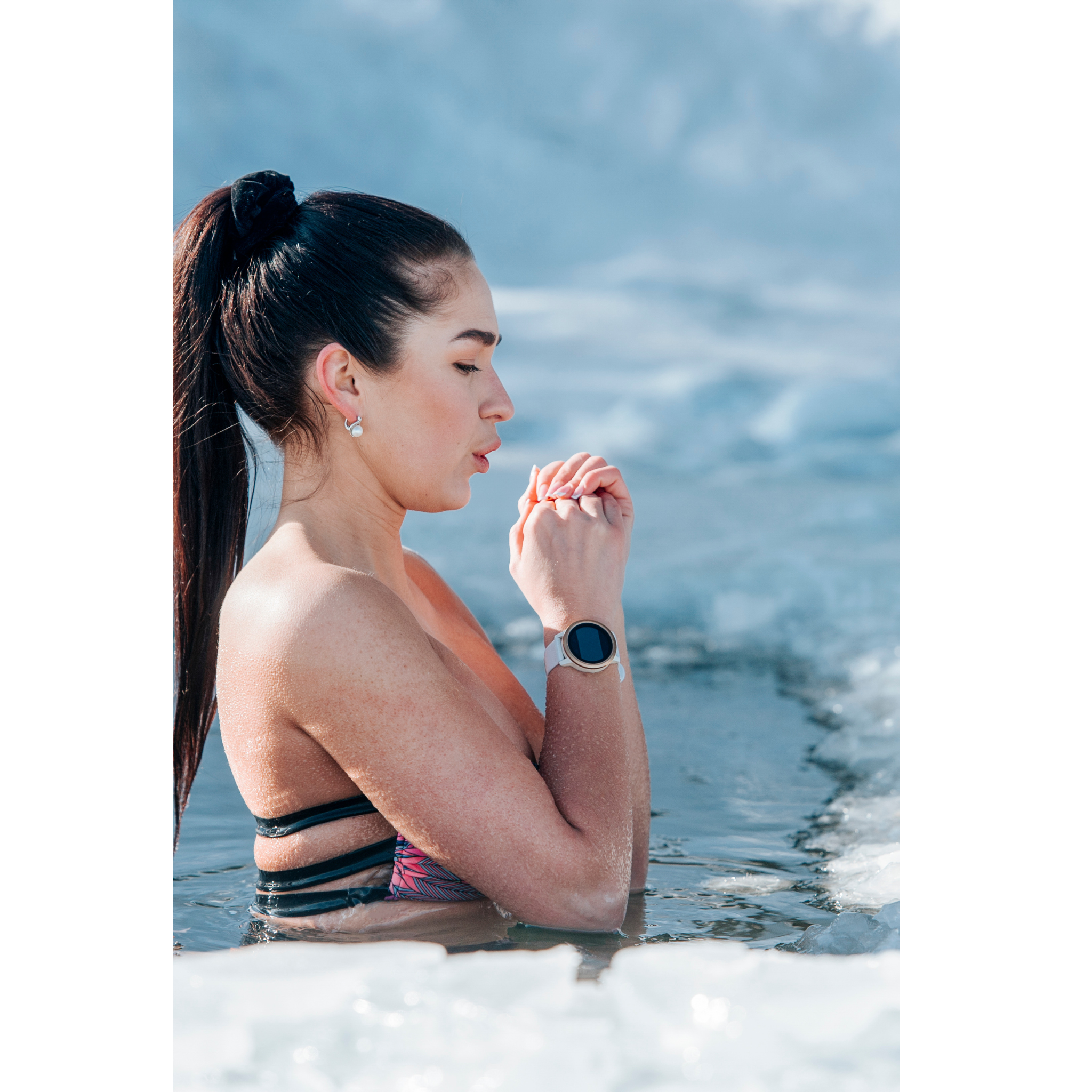 A woman with dark hair in a ponytail, wearing earrings and a fitness watch, prays or meditates in the water at a beach. ice bath