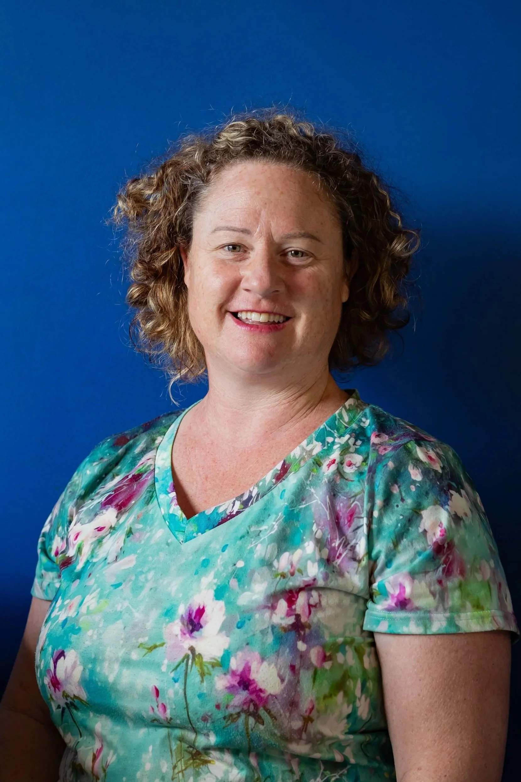 Portrait of a woman with curly hair wearing a colorful floral dress against a blue background.