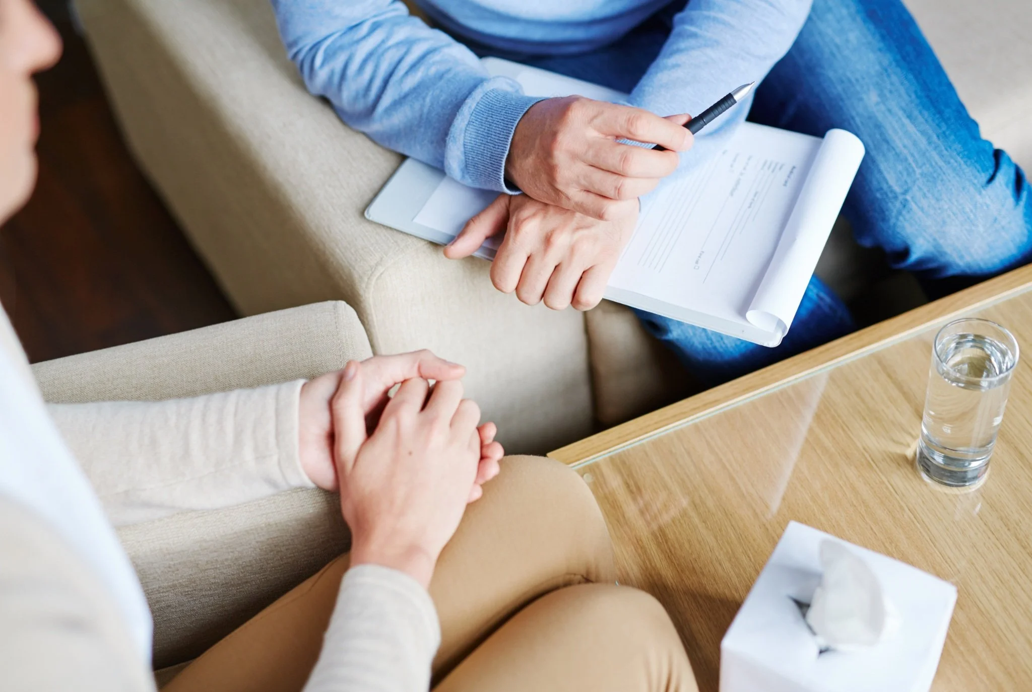A person sitting on a sofa taking notes in a notebook during a counseling session with a therapist, whose hands are visible holding a pen and a clipboard with papers. A glass of water and a tissue box are on the wooden table.