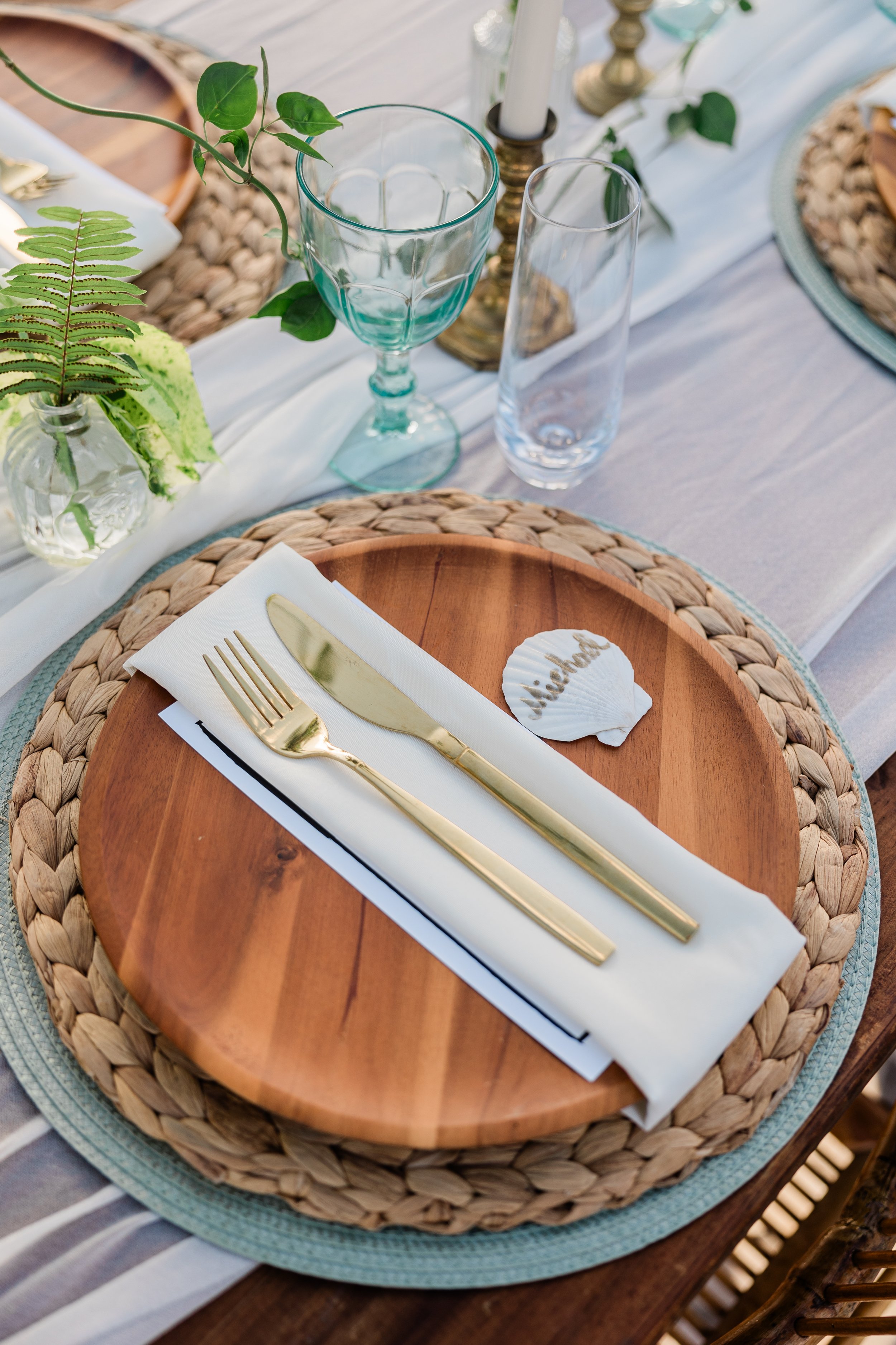 Table setting with a wooden plate, gold fork and knife on a white napkin, a seashell with the name Nicole, and various glasses and decorative plants.