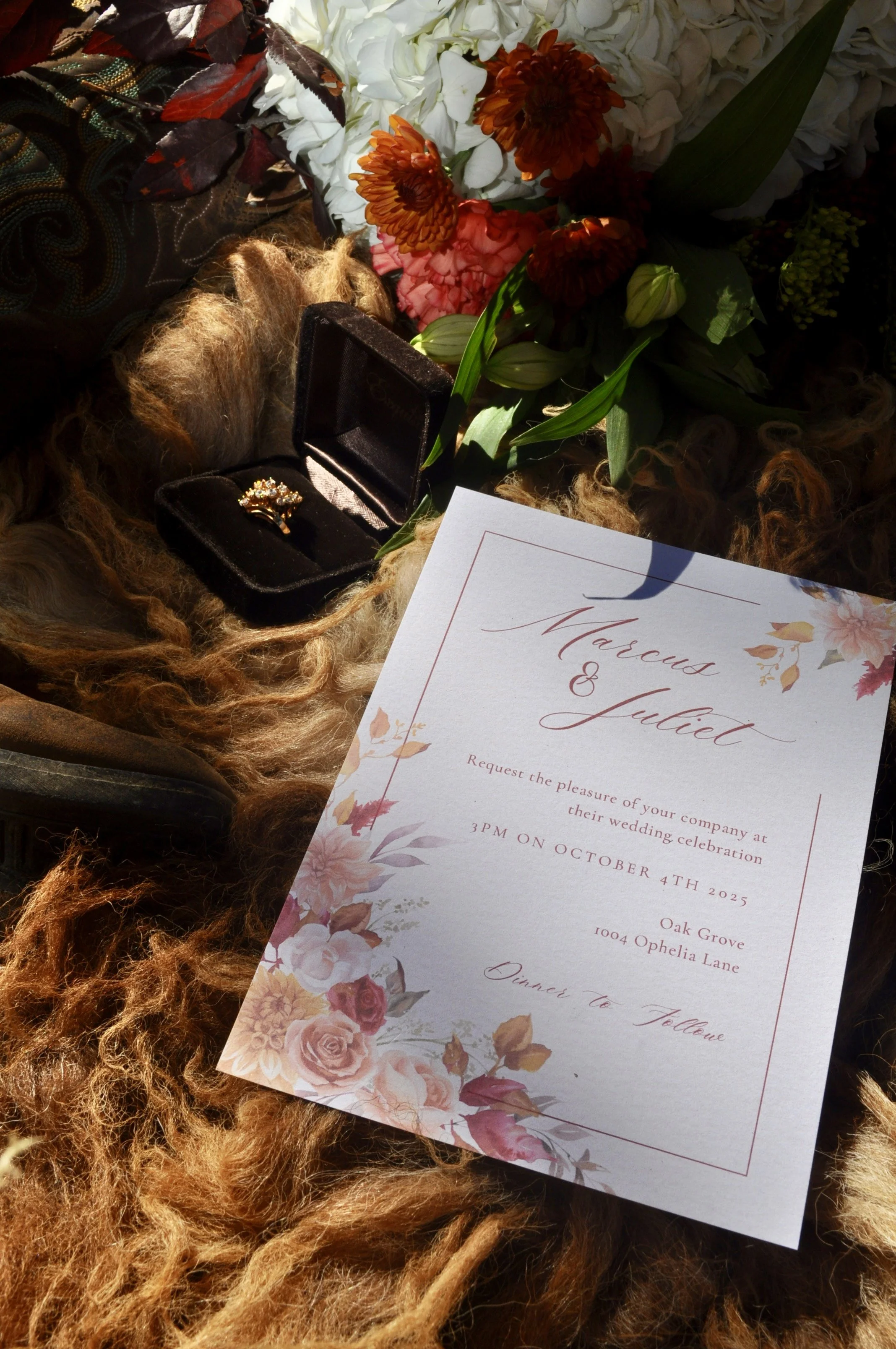 A wedding invitation card with floral design, a bouquet of flowers, and a ring in a black velvet box, all arranged on a soft textured surface.