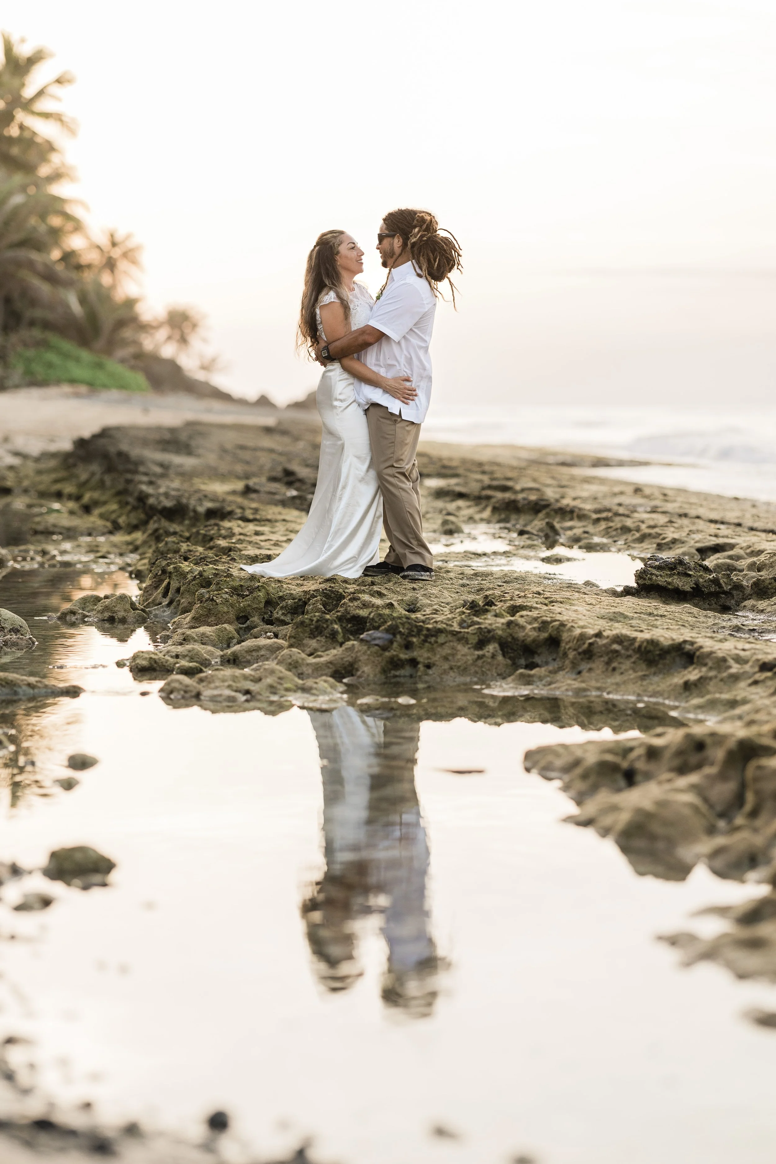 A couple stands on a rocky beach at sunset, holding each other and looking into each other's eyes, with the ocean and palm trees in the background.