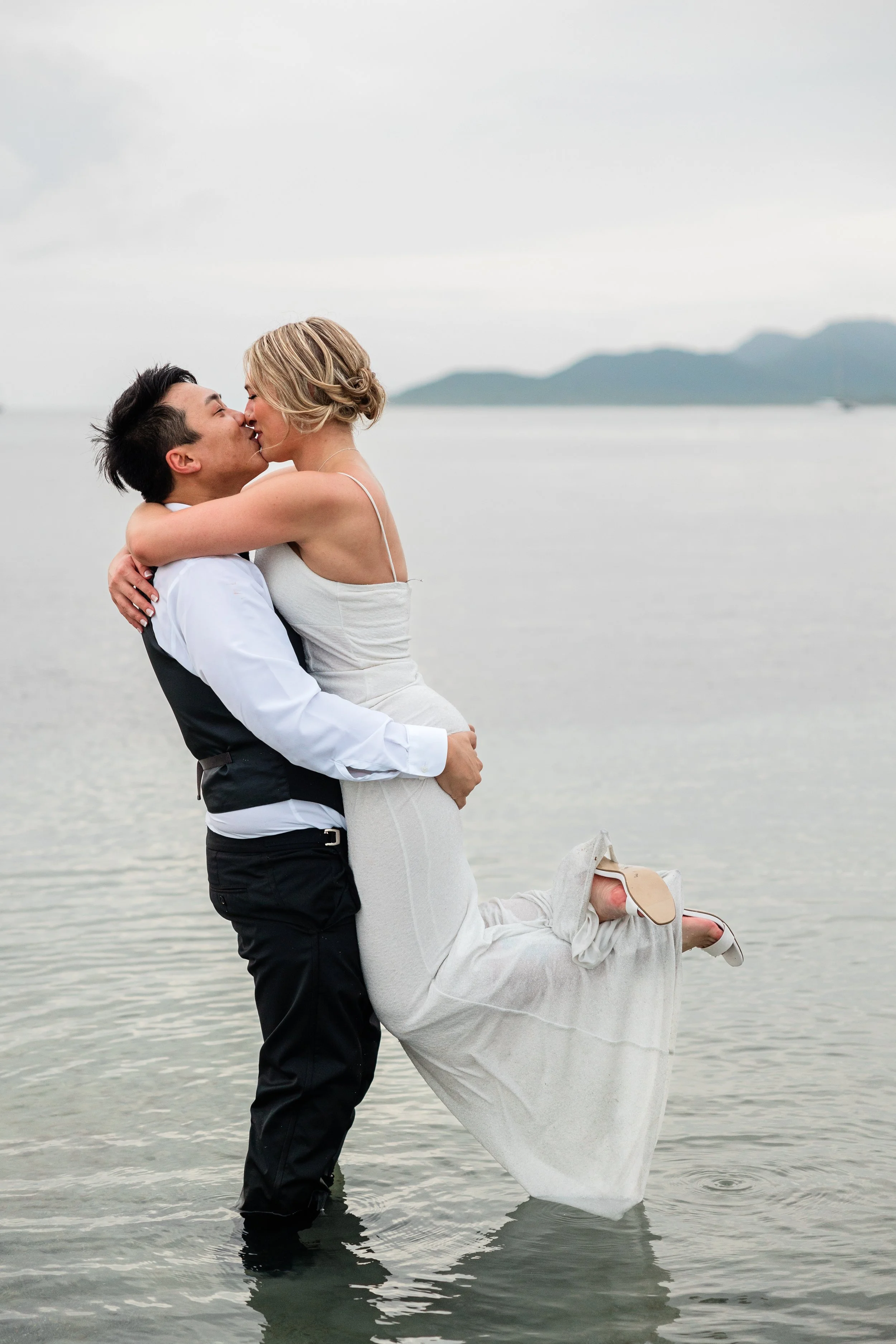 A couple dressed in wedding attire stands in water, kissing, with mountains in the background.