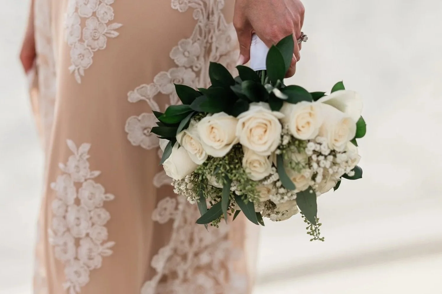 A bride in wedding dress with lace floral embroidery on the side holds a bouquet of white roses, greenery, and baby's breath.