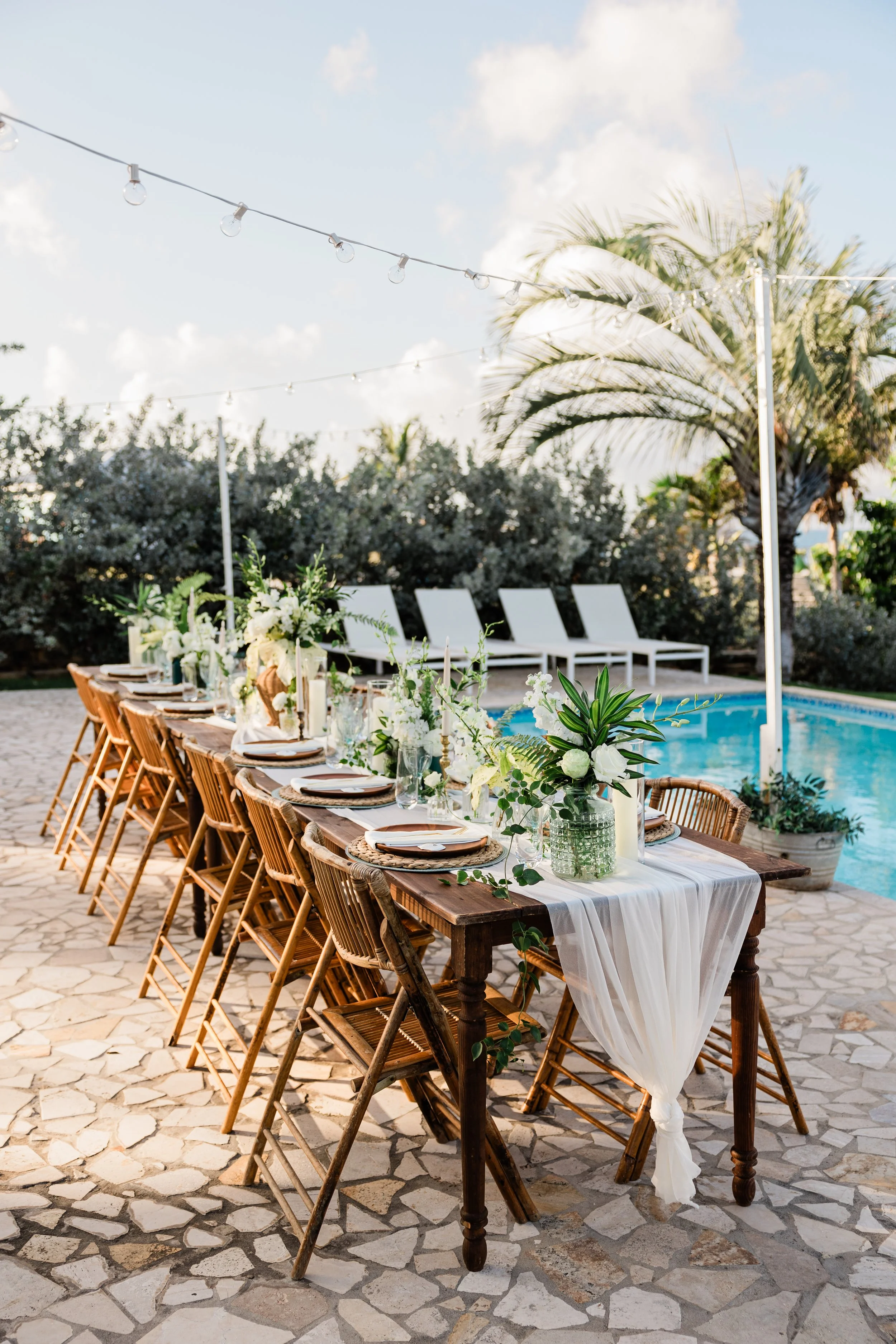 Outdoor dining table set for a celebration by a poolside, decorated with white flowers and candles, surrounded by lounge chairs, under string lights and palm trees.