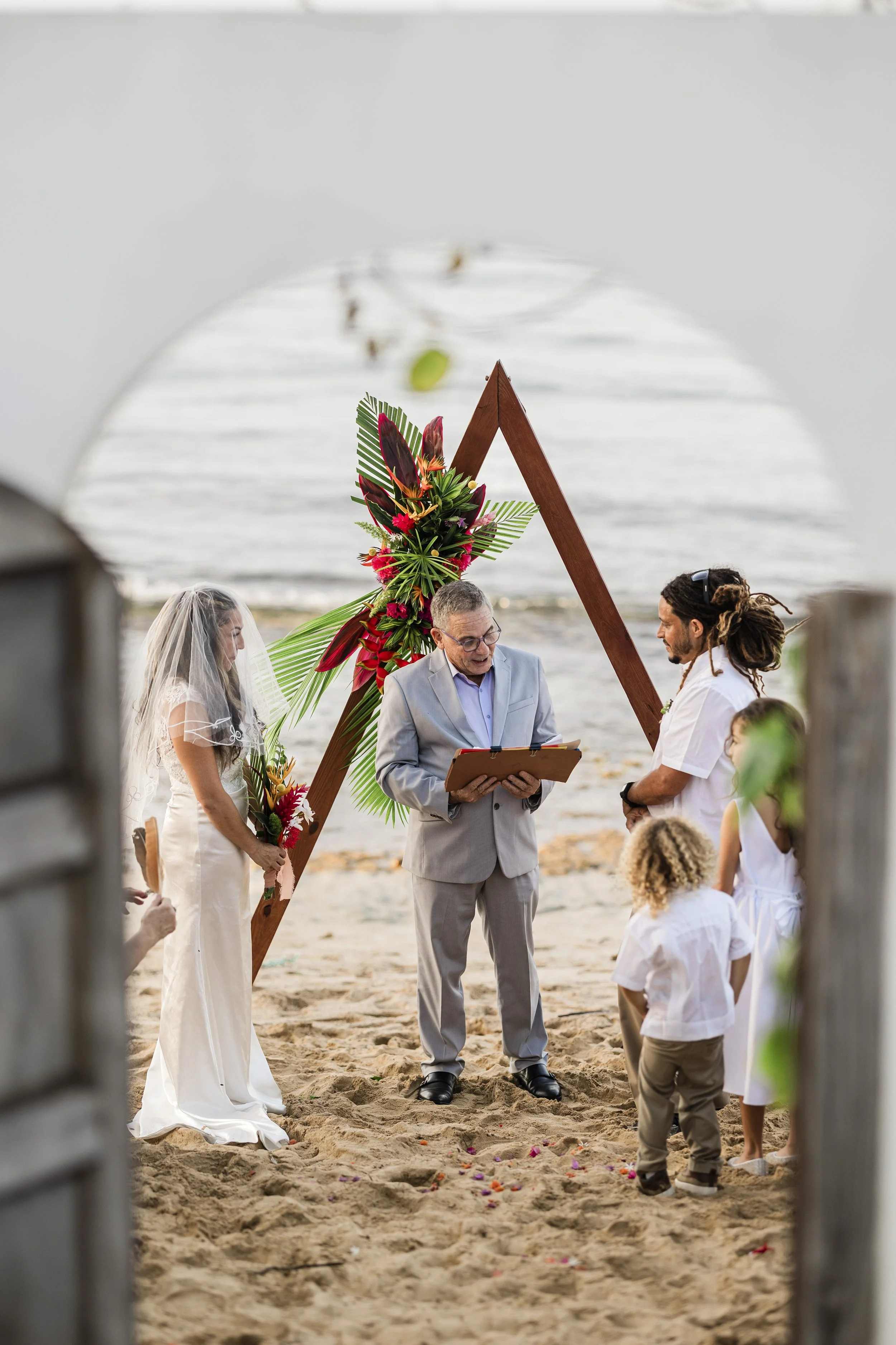 A beach wedding ceremony with a priest officiating, a bride in a white dress and veil, a groom with dreadlocks, and children, with a wooden arch decorated with tropical flowers and greenery, near the ocean.