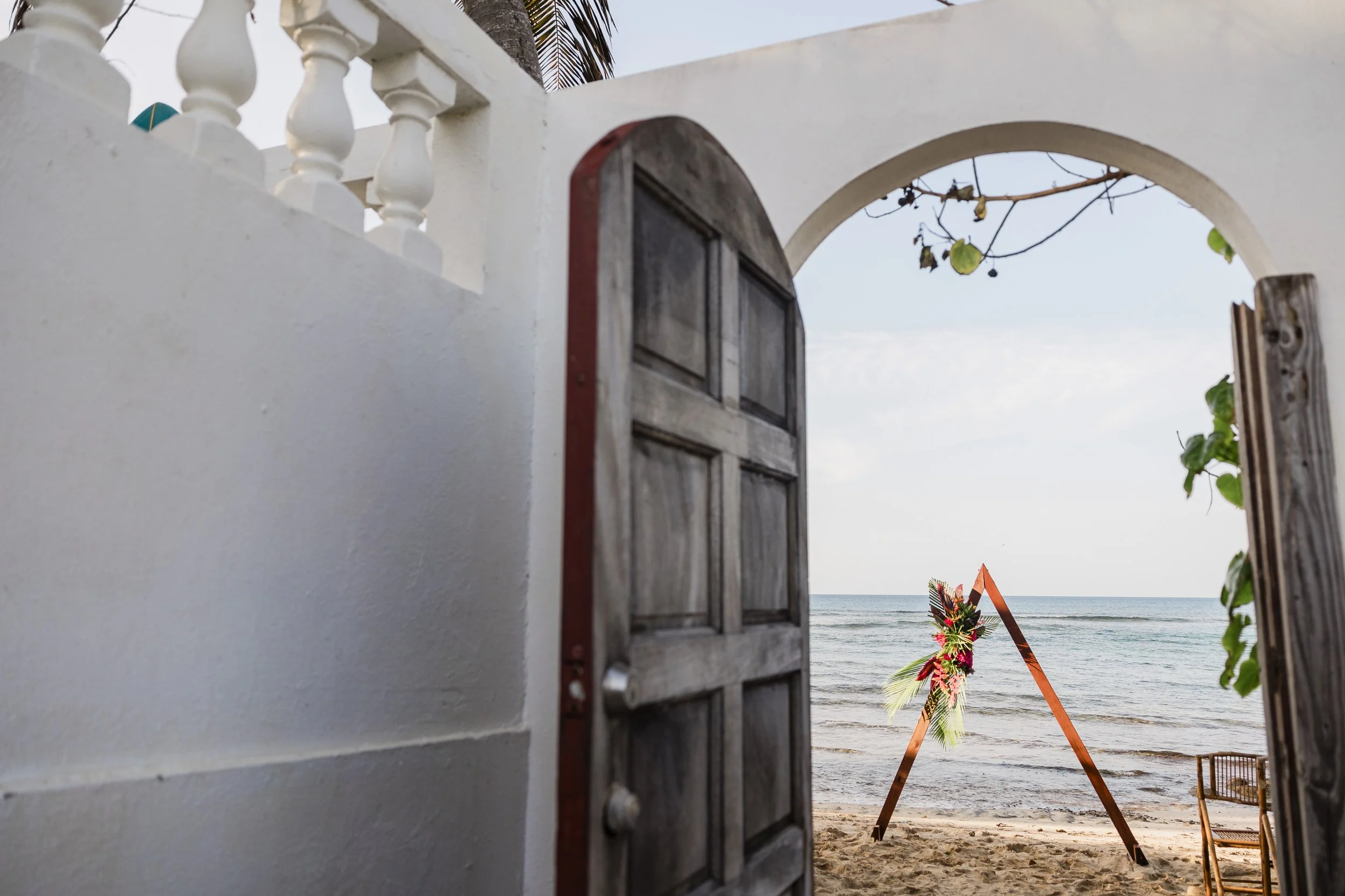 Beach wedding setup with a floral arch, view through an open wooden gate, ocean in the background, sandy shore, and a white wall with decorative balusters.