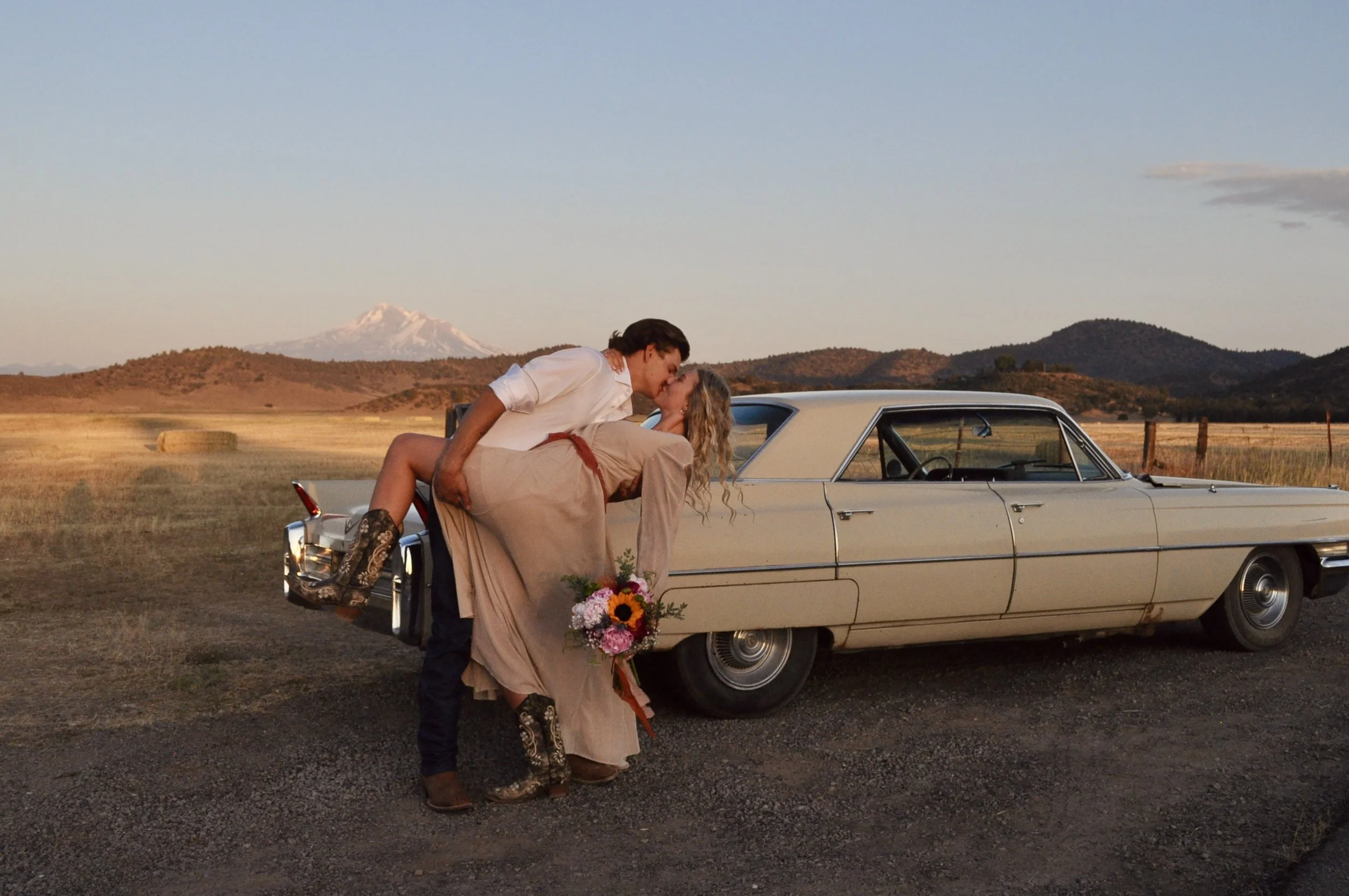 A couple kissing next to a vintage car in a rural landscape at sunset, with mountains in the background.