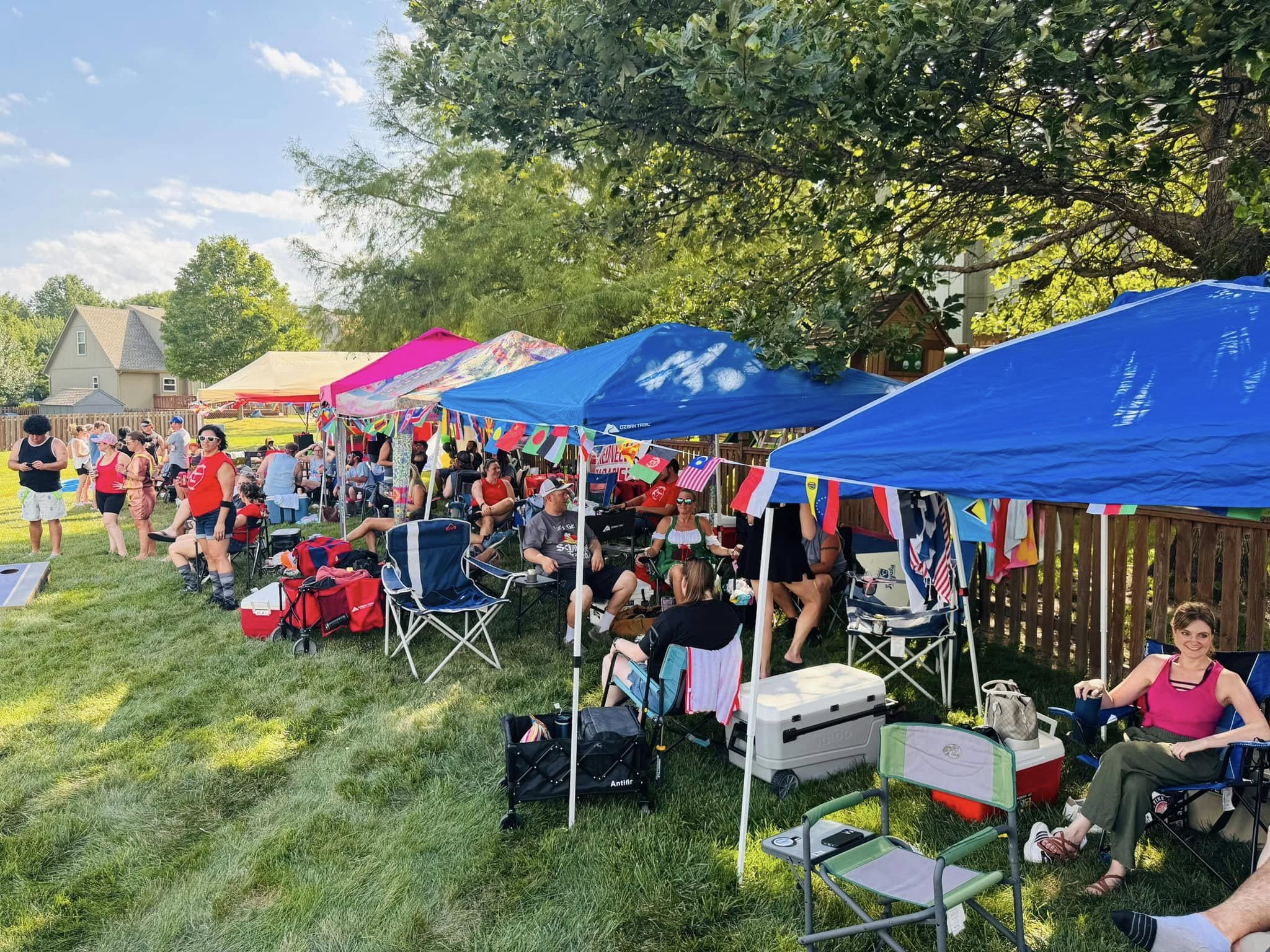 People gathered in a backyard with tents and chairs, celebrating outdoors on a sunny day with green grass and trees.
