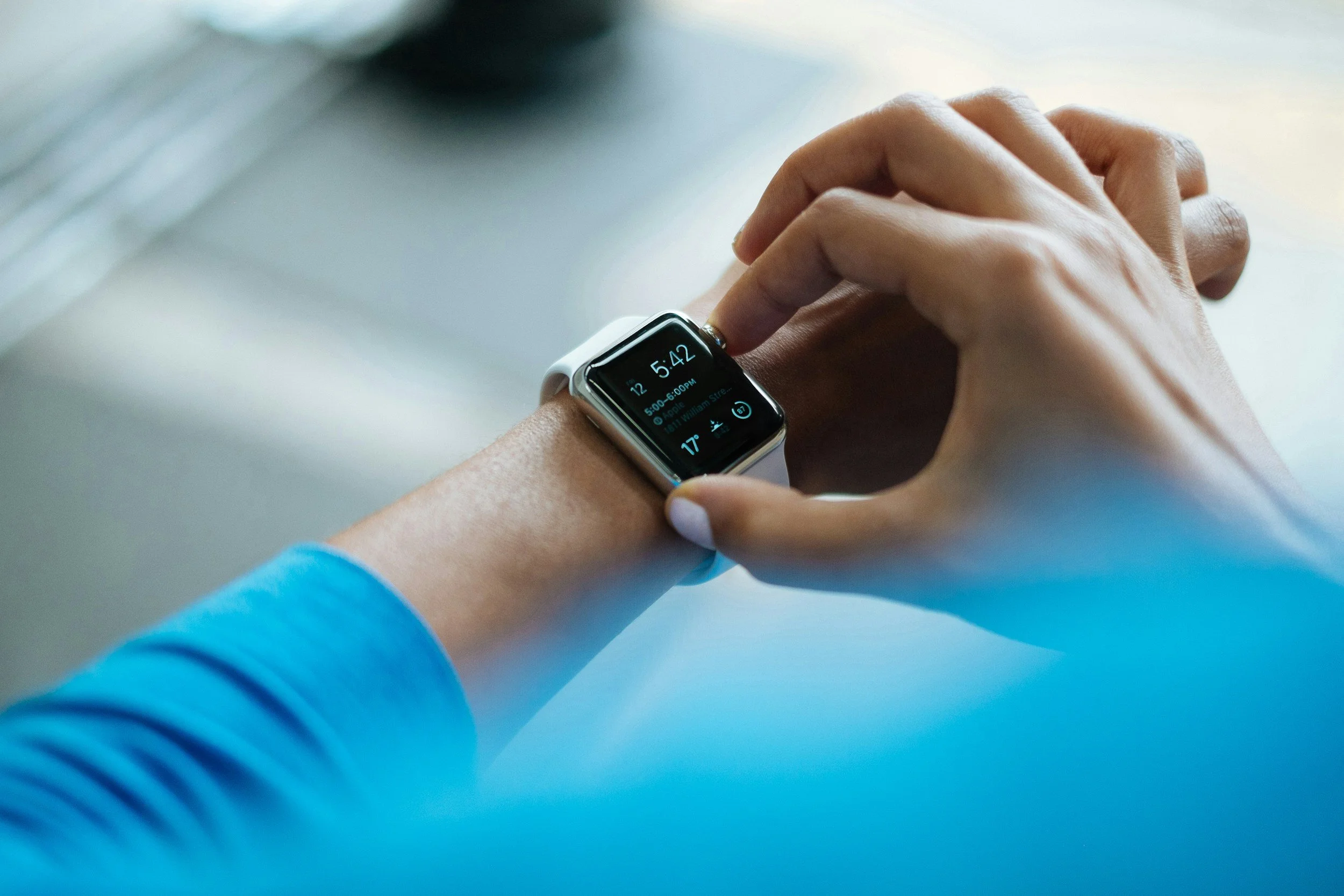 A person wearing a white smartwatch with a black screen displaying the time 5:42, date, and weather information, adjusting the watch with their right hand.