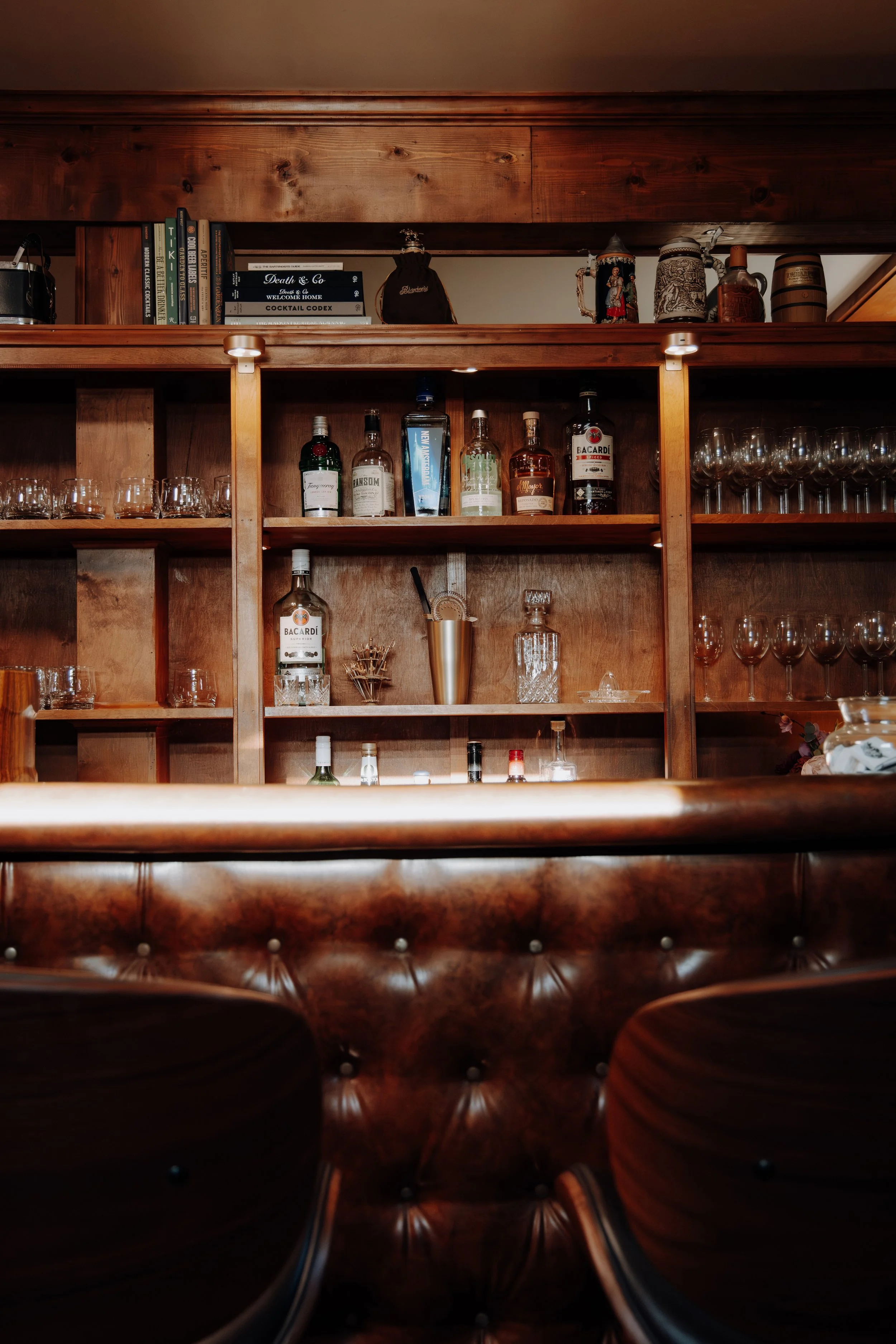 Wooden bar shelf with various liquor bottles, glassware, books, and decorative items in a dimly lit setting.
