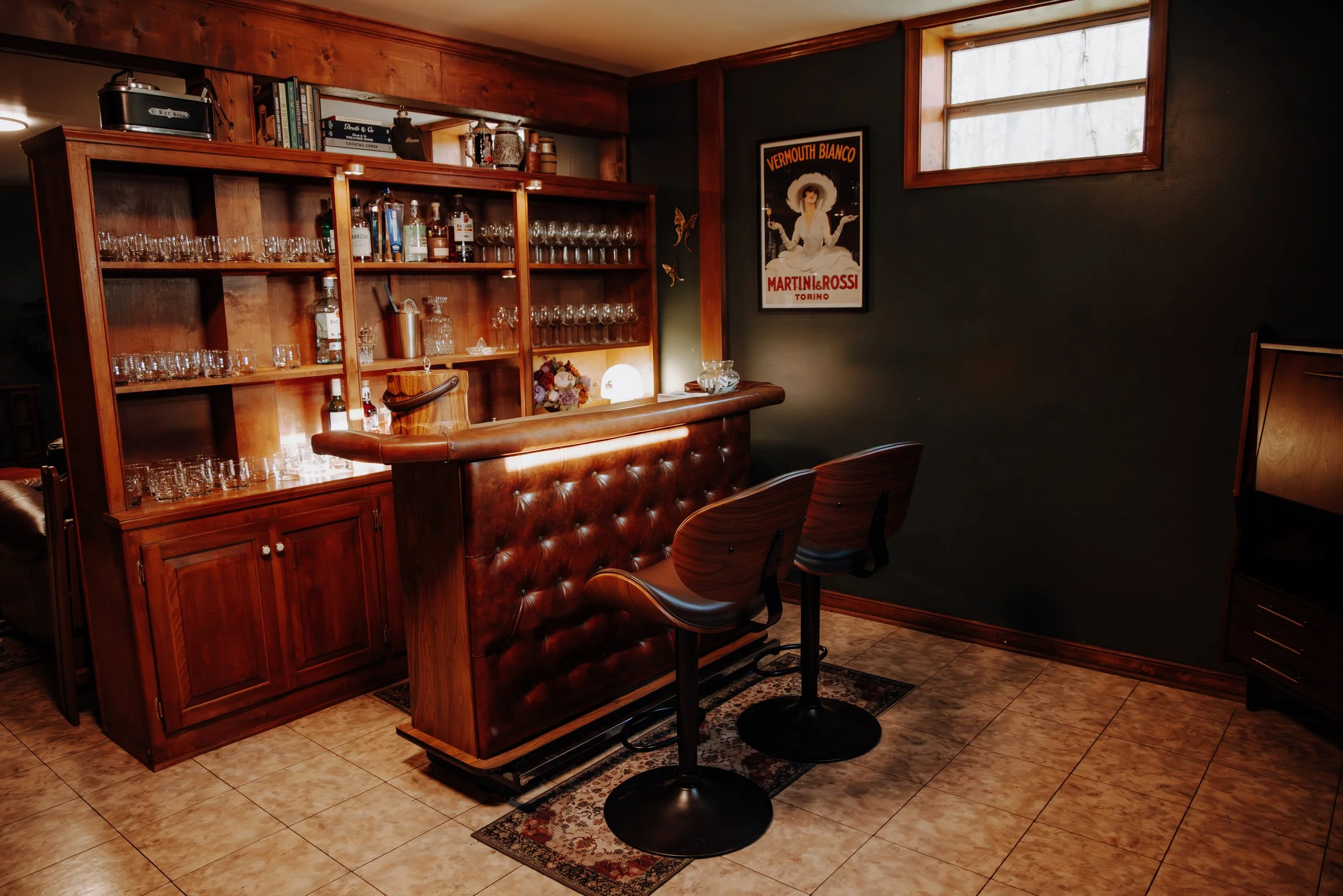 A cozy home bar with two wooden barstools in front of a polished wood bar counter. Behind the bar, a built-in wooden shelf holds various liquor bottles and glassware. The wall to the right has a vintage poster and there is a window near the ceiling allowing natural light into the room.