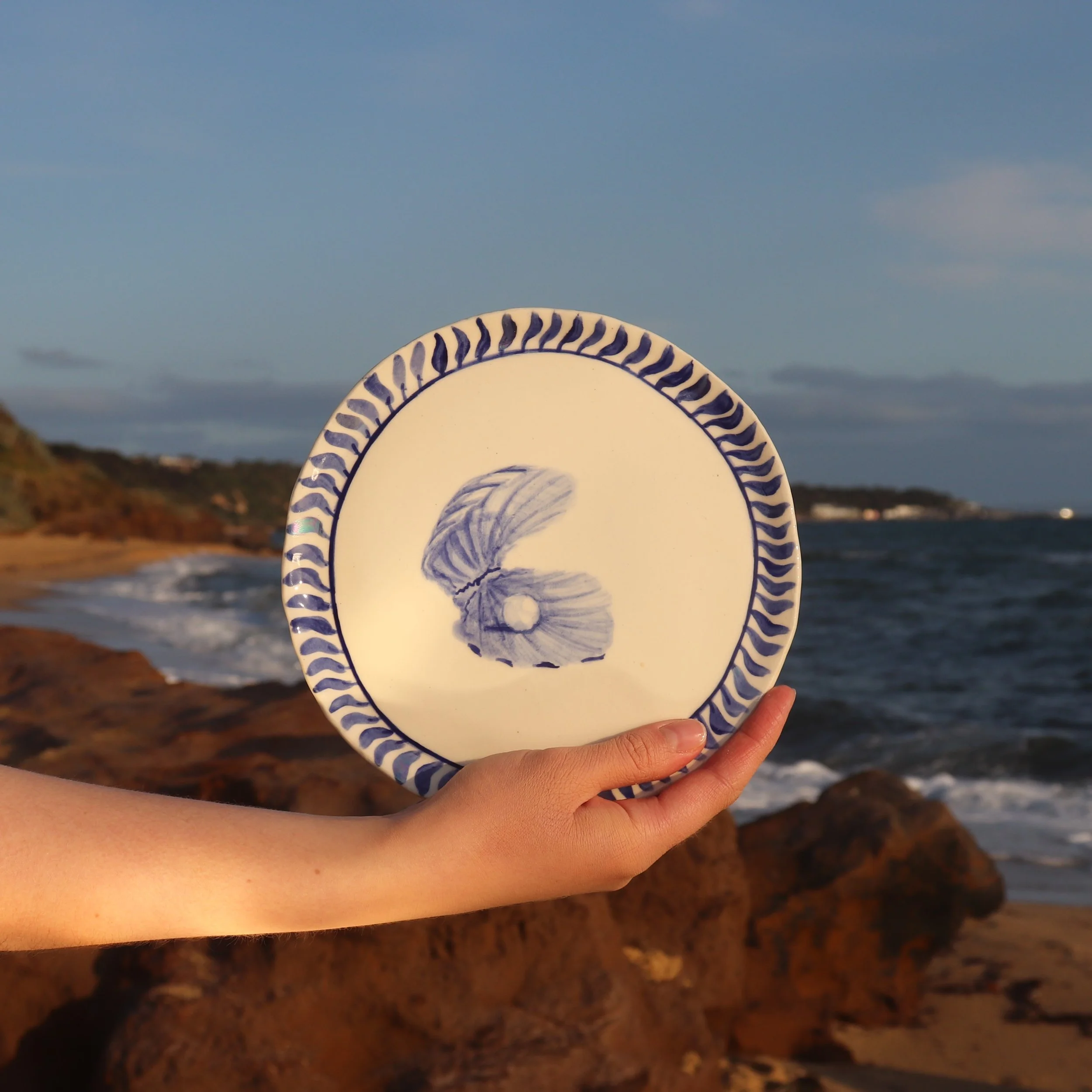 Person holding a decorative plate with a blue and white design, featuring a shell motif, against a beach background with rocks, ocean, and sky.