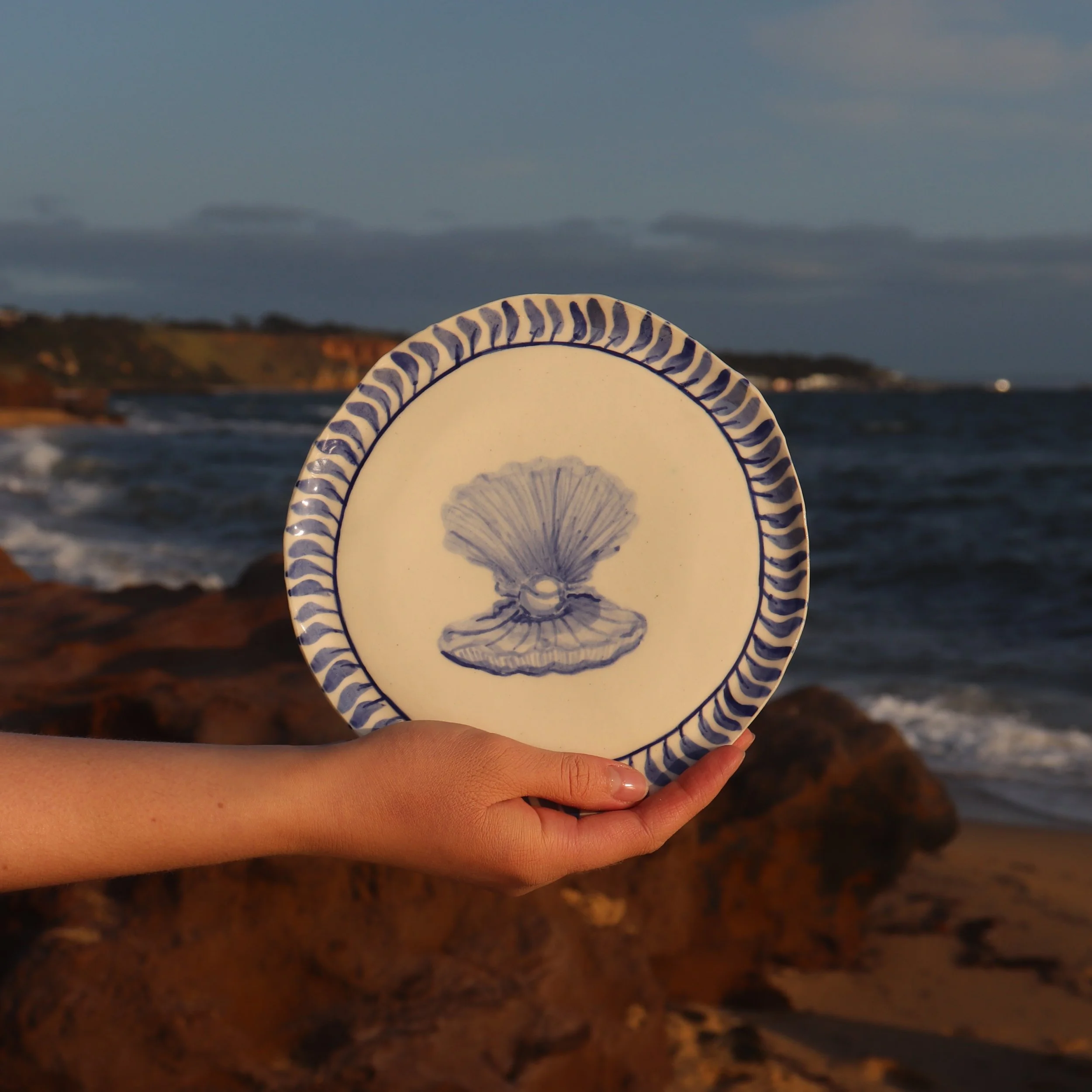 A hand holding a decorative plate with a blue and white shell design in front of a beach and ocean scene during sunset.