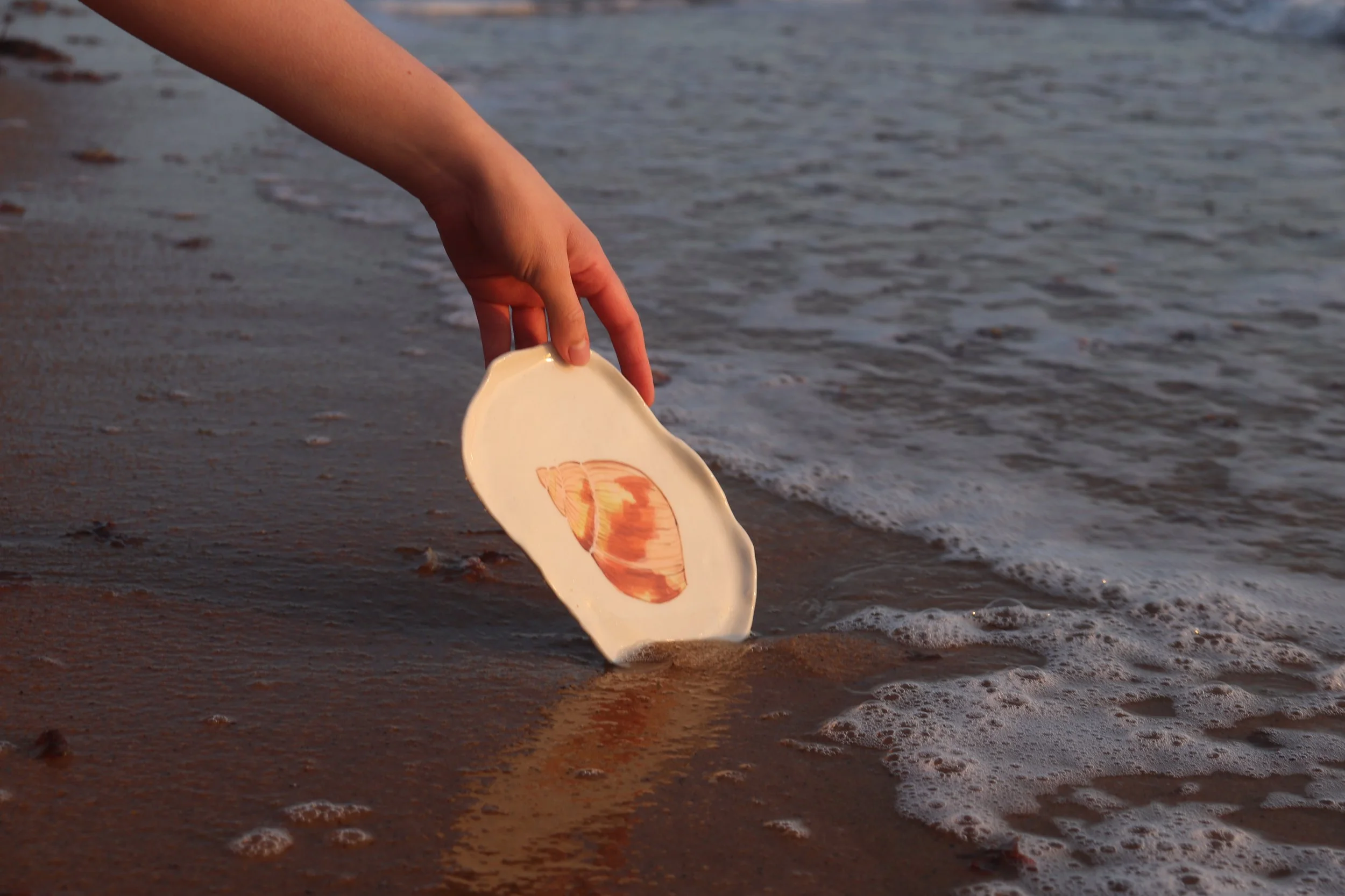 Person's hand holding a ceramic shell-shaped dish with an image of a real seashell, near the shoreline with foamy waves.