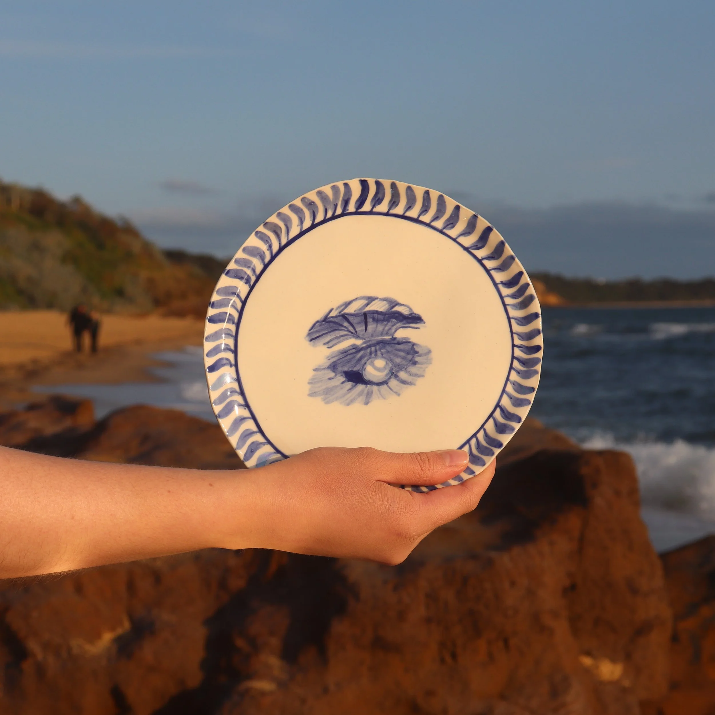 Person holding a decorative ceramic plate with a blue and white seashell design in front of a beach scene with rocks, ocean waves, a sandy shore, and a horse in the distance during sunset.