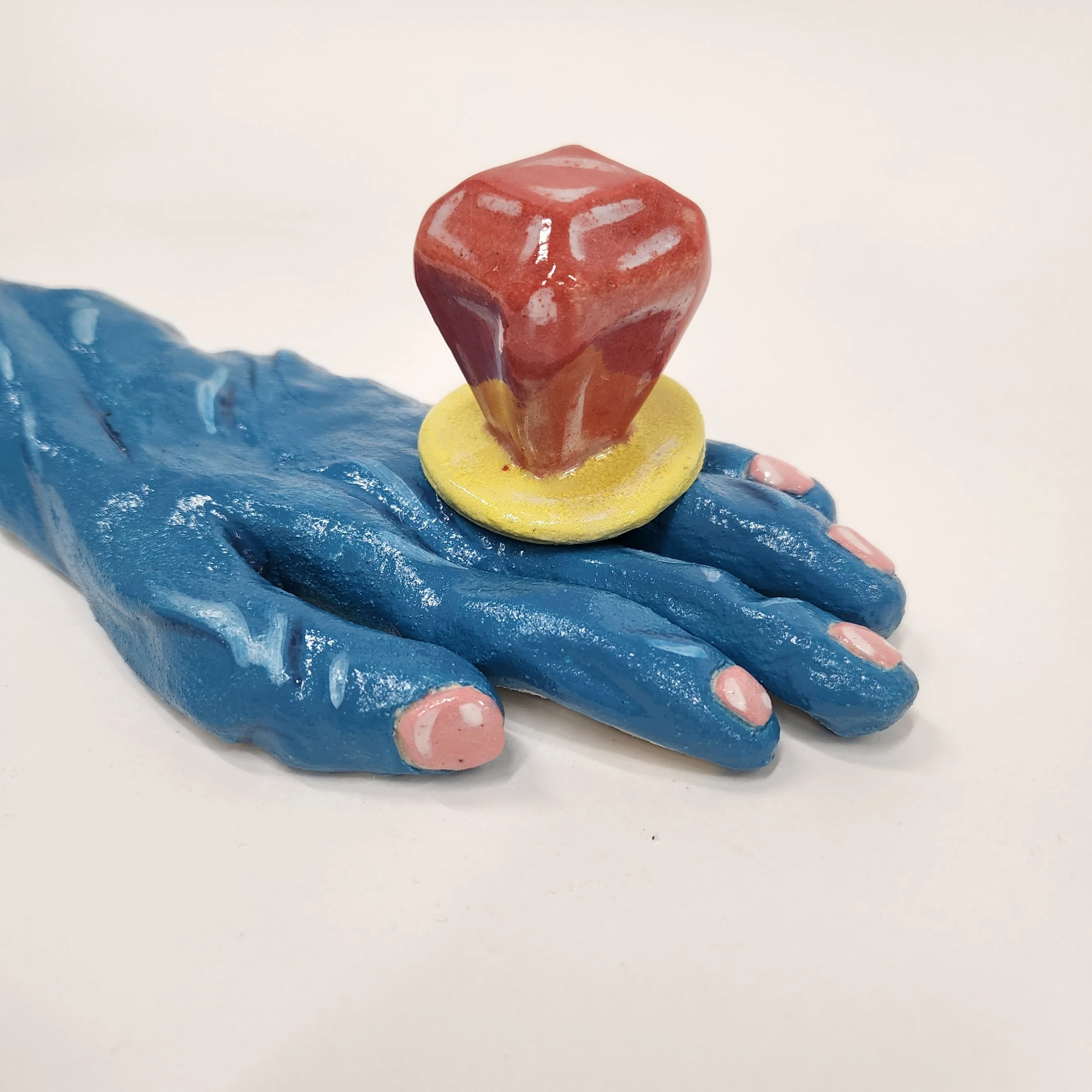 A blue ceramic hand sculpture with a large yellow ring with a red gemstone on top.