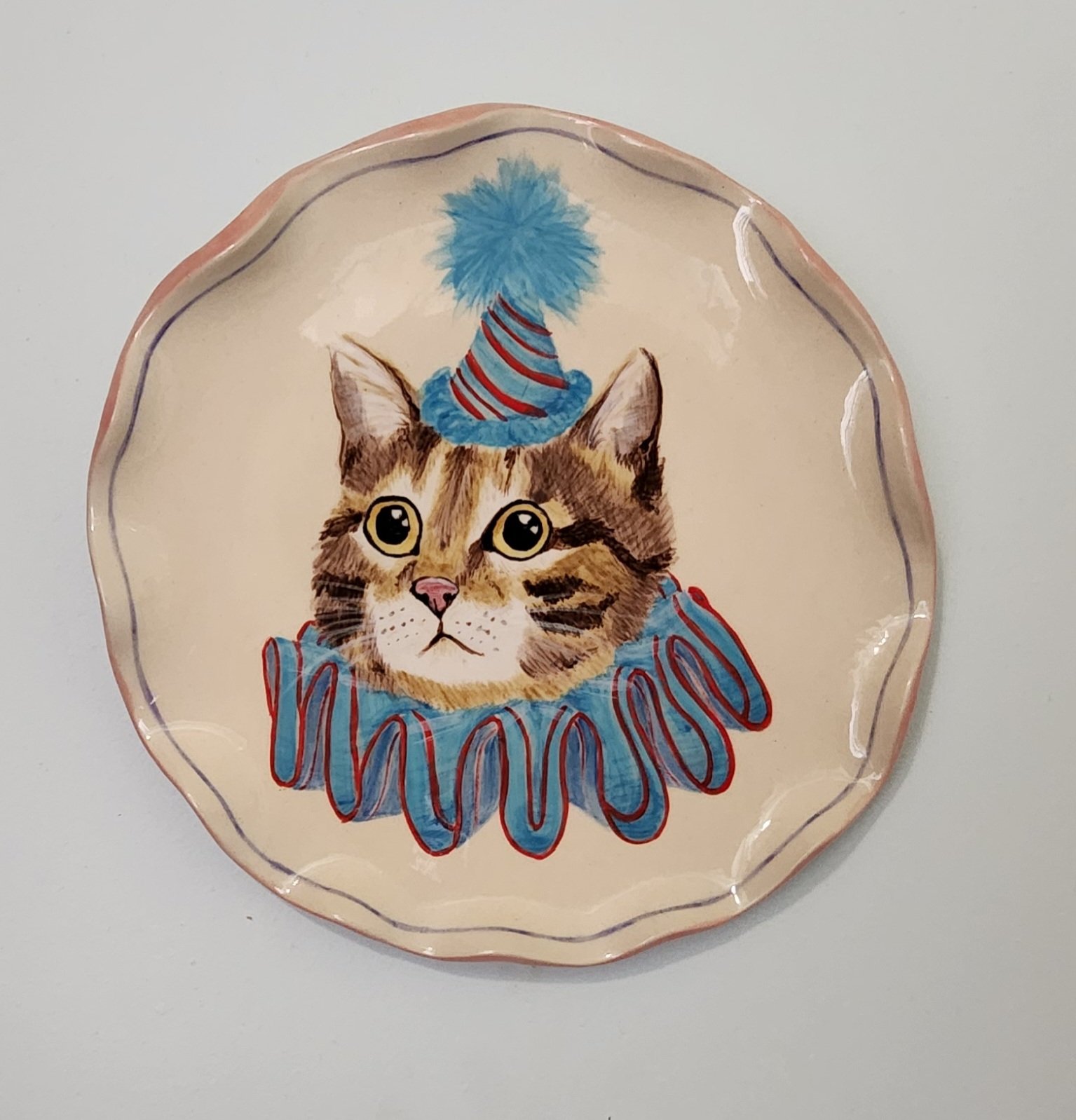 Decorative plate featuring a realistic illustration of a cat wearing a clown costume, with a blue and red hat with a pom-pom and a ruffled collar.