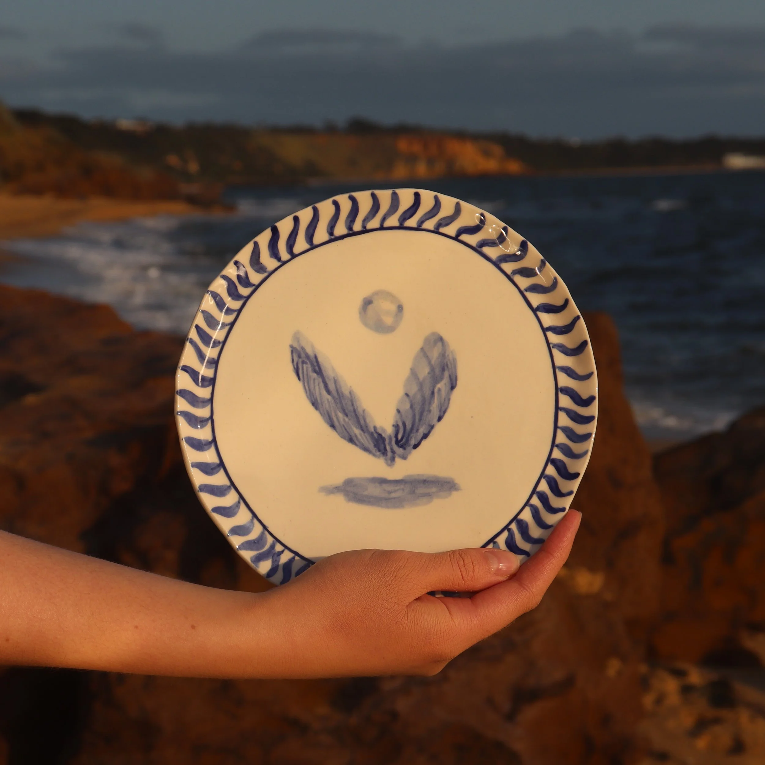 A person holding a decorative ceramic plate with blue and white design, featuring a hand-painted shell, against a coastal background with cliffs and ocean during sunset.