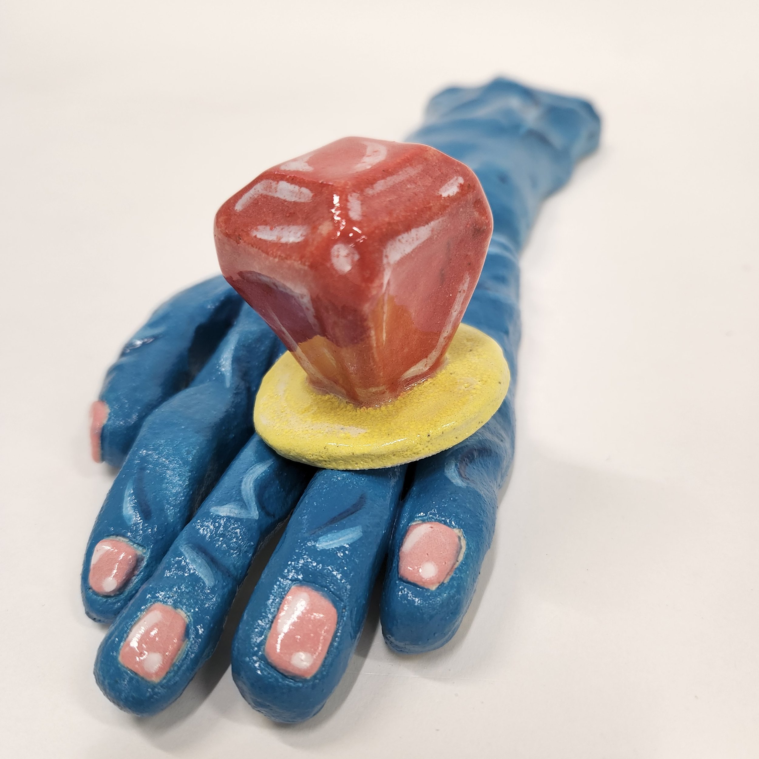 A blue ceramic hand sculpture with a large yellow ring with a red gemstone on top.