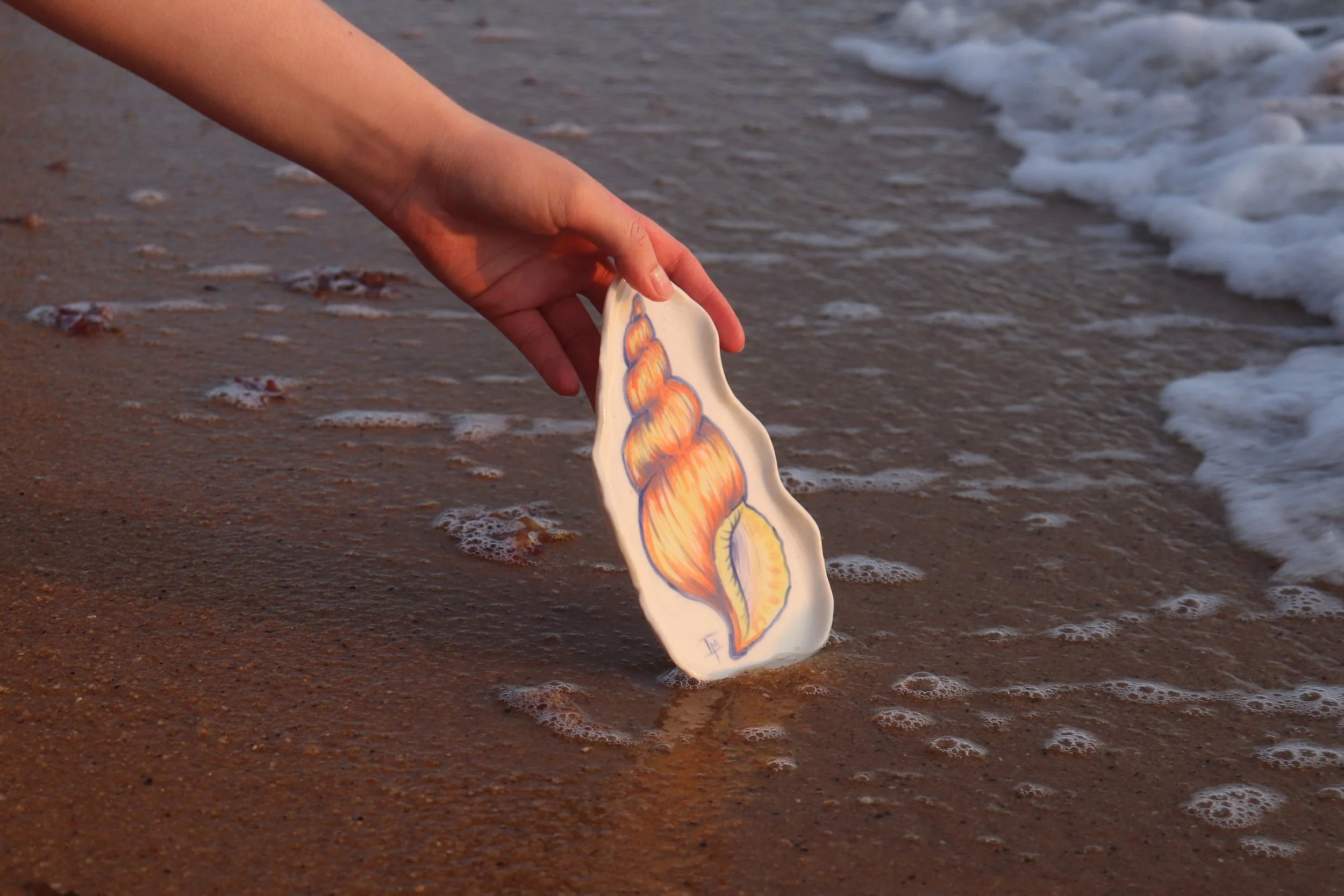 Person's hand holding a decorated ceramic plate with a colorful seashell drawing, as it interacts with the shoreline and shallow waves on the beach.
