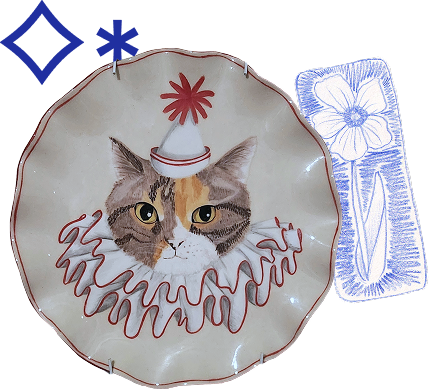 Decorative porcelain plate with a cat wearing a clown hat and curly ruffle, alongside a drawing with a flower design, set against a blue background.