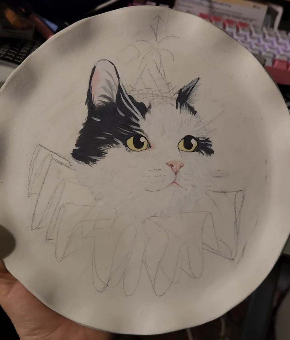 Partially finished painted ceramic plate with a realistic depiction of a black and white cat's face and yellow eyes, surrounded by faint sketches of floral and leaf patterns.