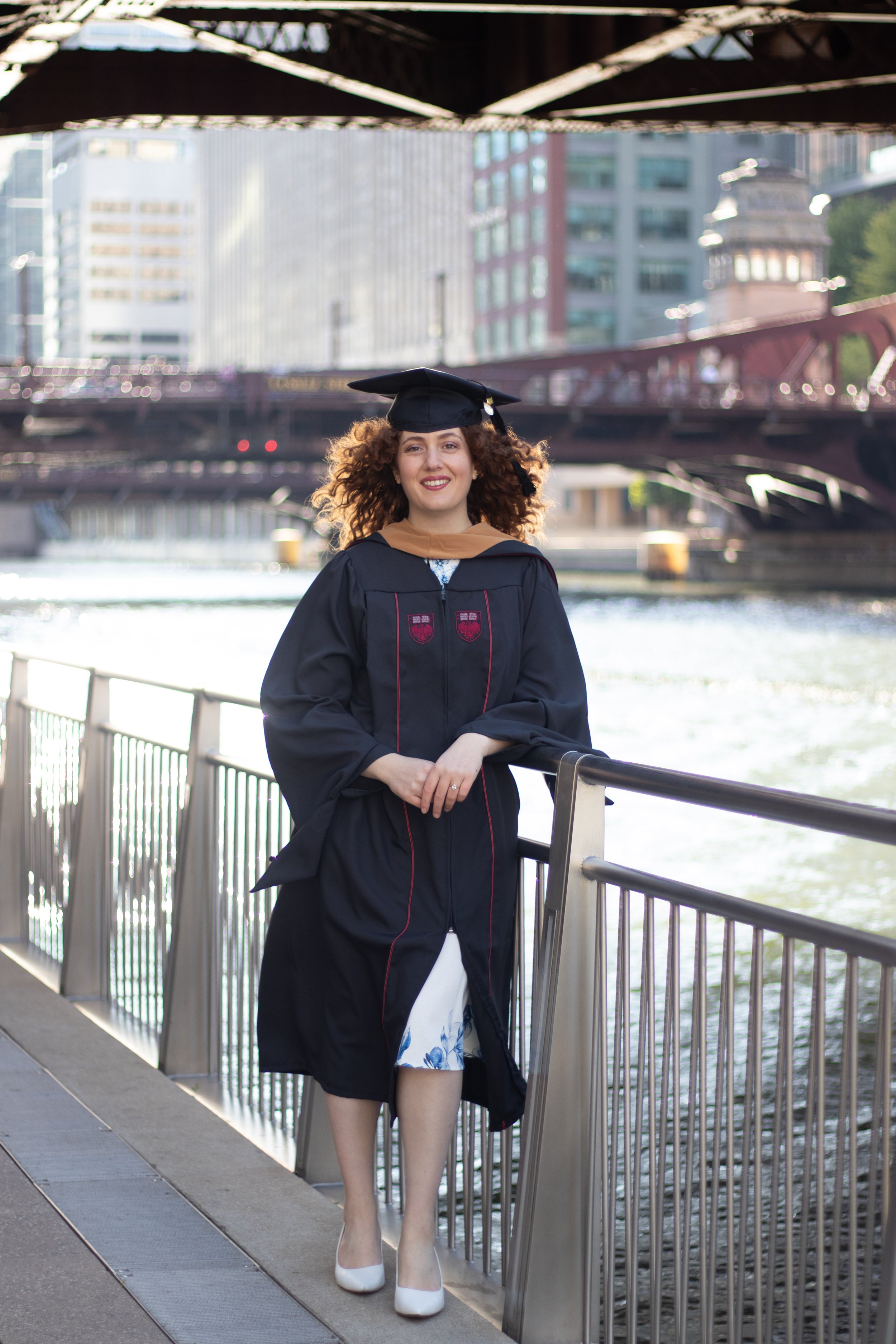 A woman in graduation attire, including a cap and gown, standing by a riverside with city buildings and a bridge in the background.