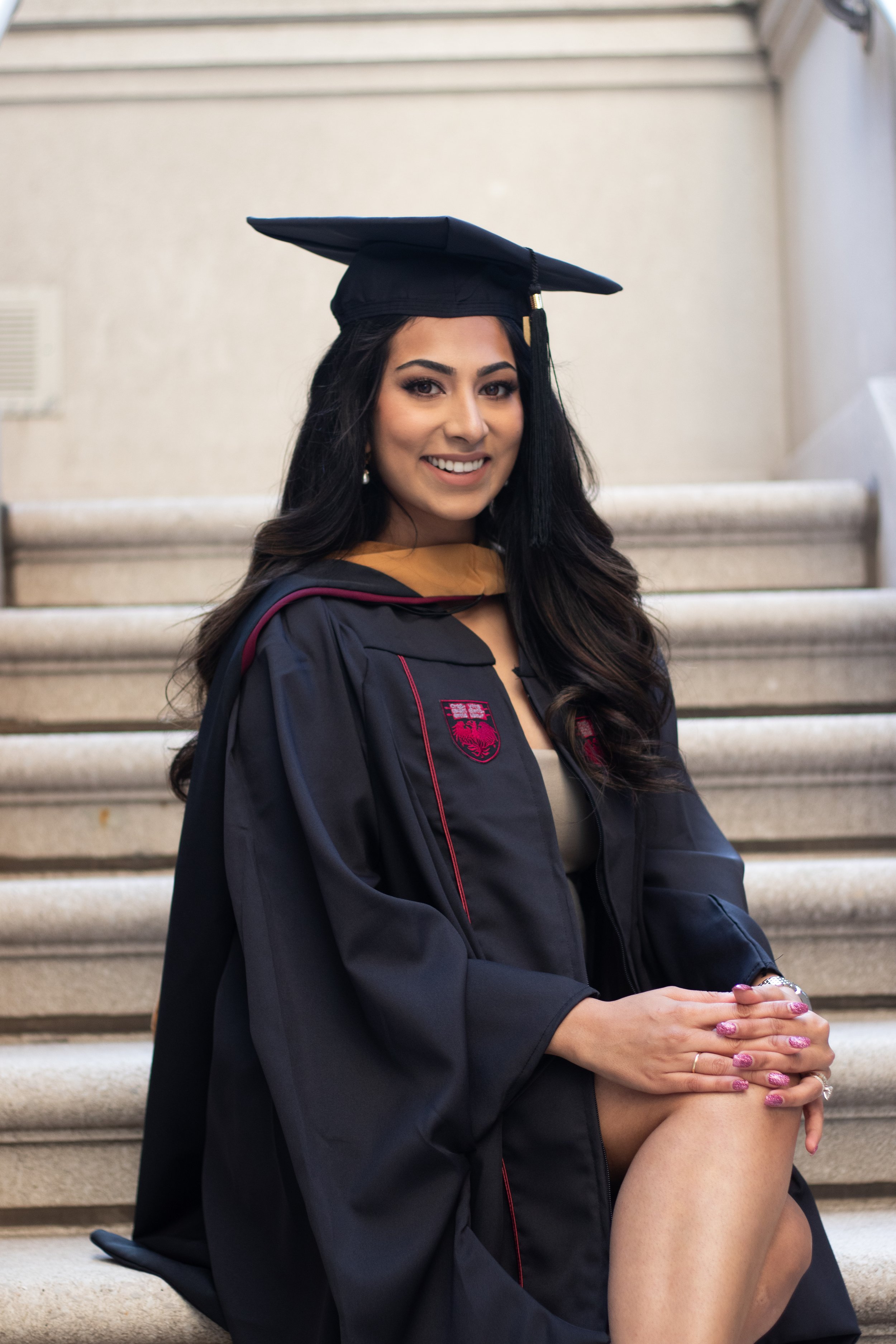 A young woman in a graduation cap and gown sitting on steps, smiling at the camera.