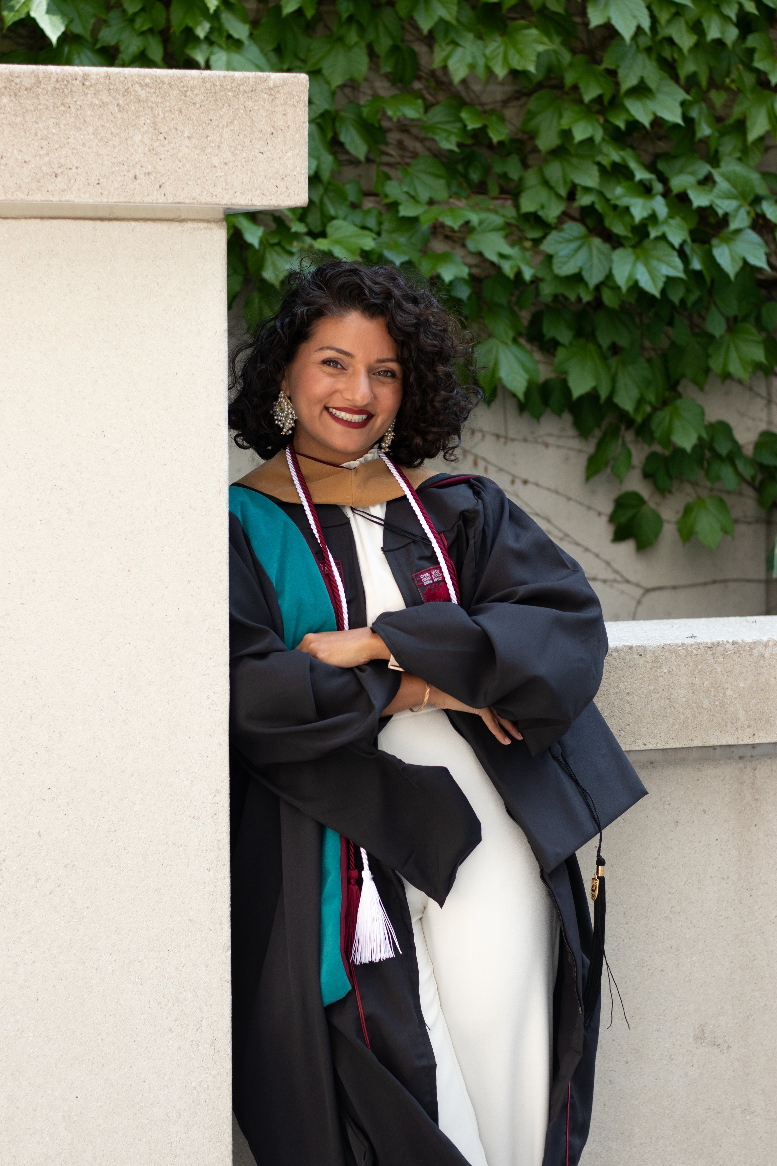 A woman in graduation gown and stole standing outdoors near a wall with green ivy, smiling with arms crossed.