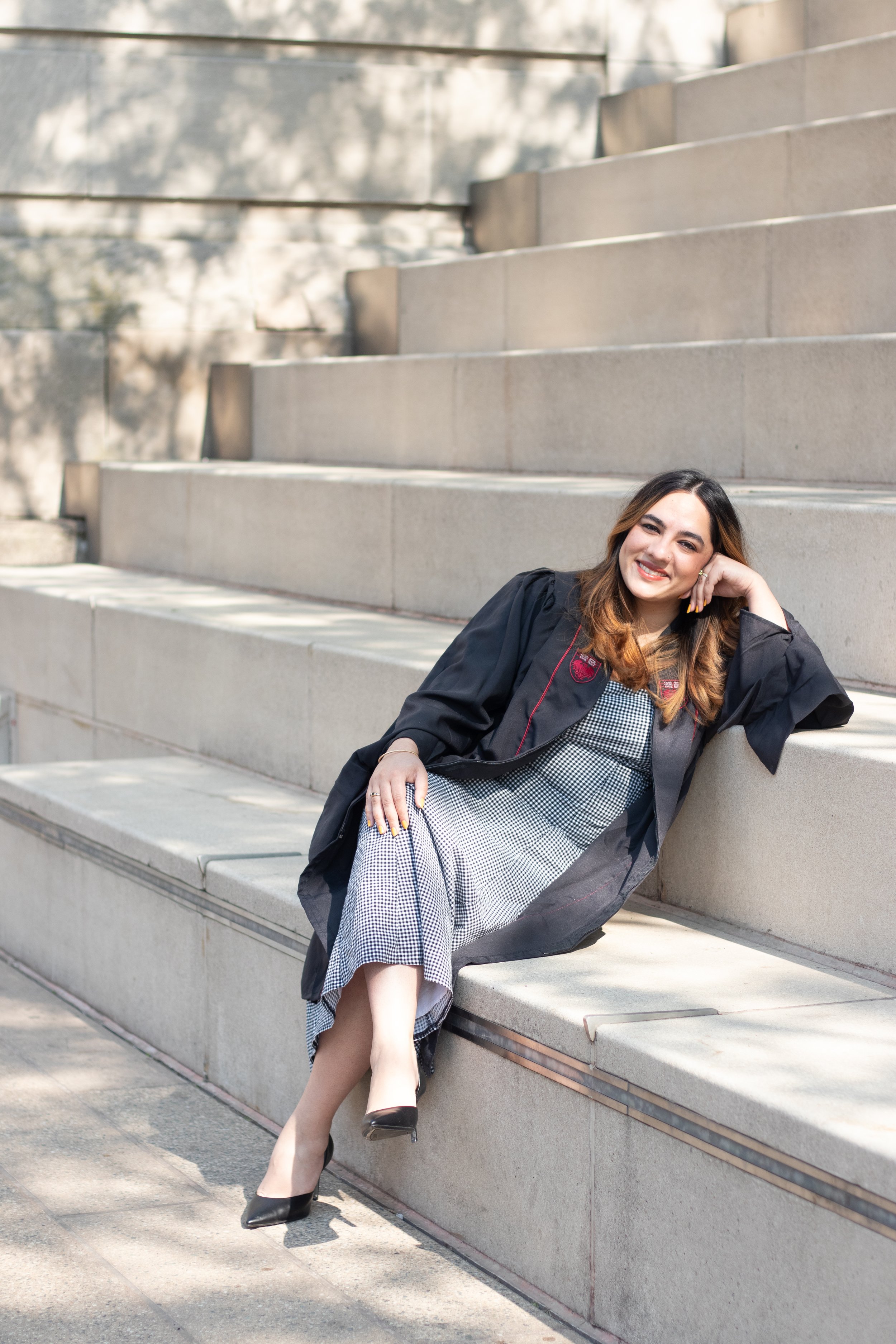 A woman wearing a graduation gown sitting on outdoor steps, smiling and resting her head on her hand.