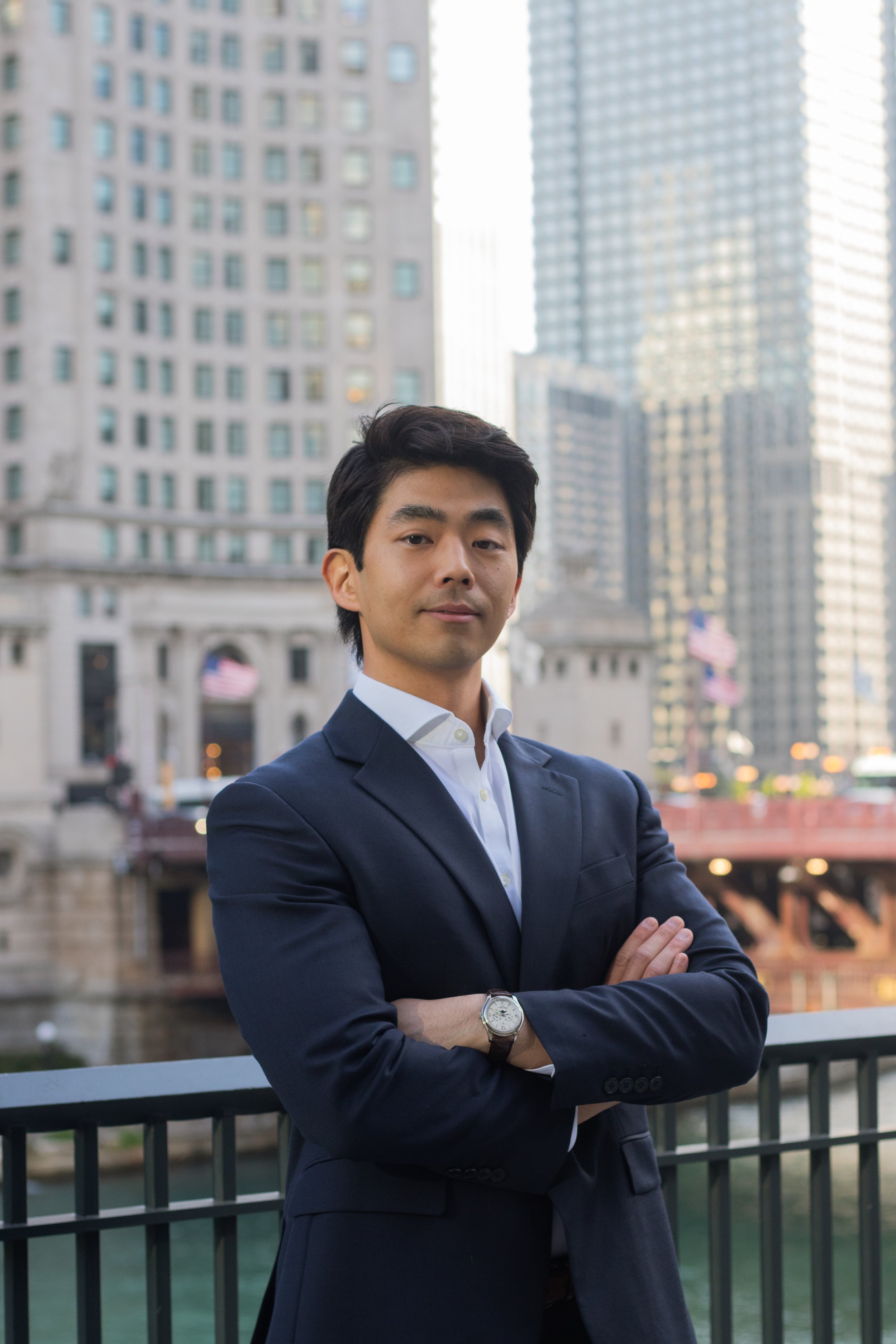A confident man in a navy suit and white shirt with crossed arms stands outdoors in an urban cityscape with tall buildings and American flags in the background.