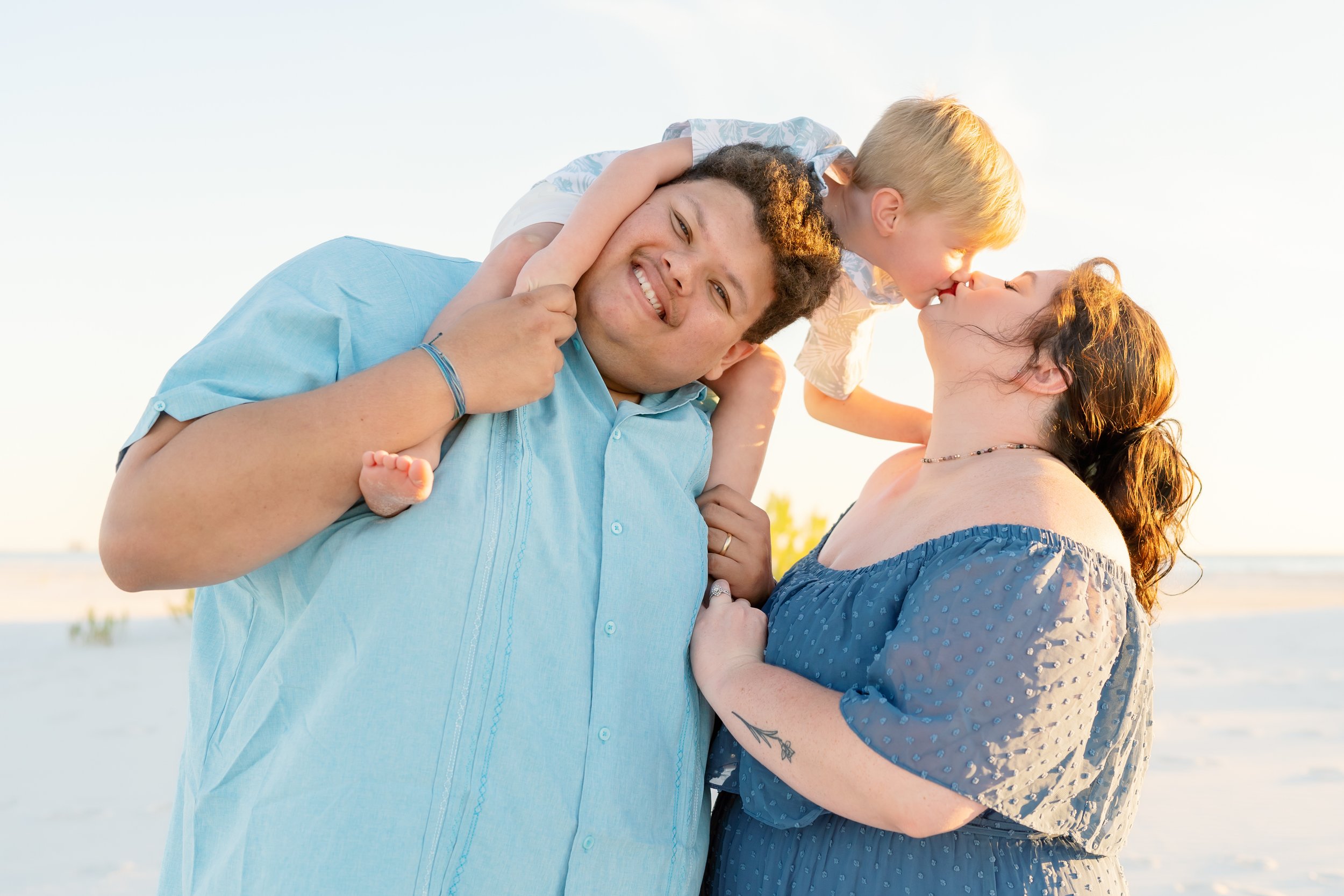 Family enjoying beach time, with child on father's shoulders and mother kissing child.