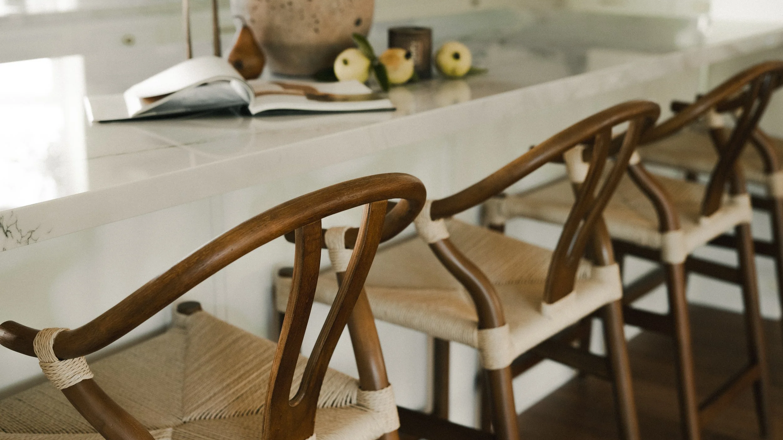 Wooden dining chairs with woven seats lined up along a white marble kitchen island with an open book and some decorative fruit in the background.