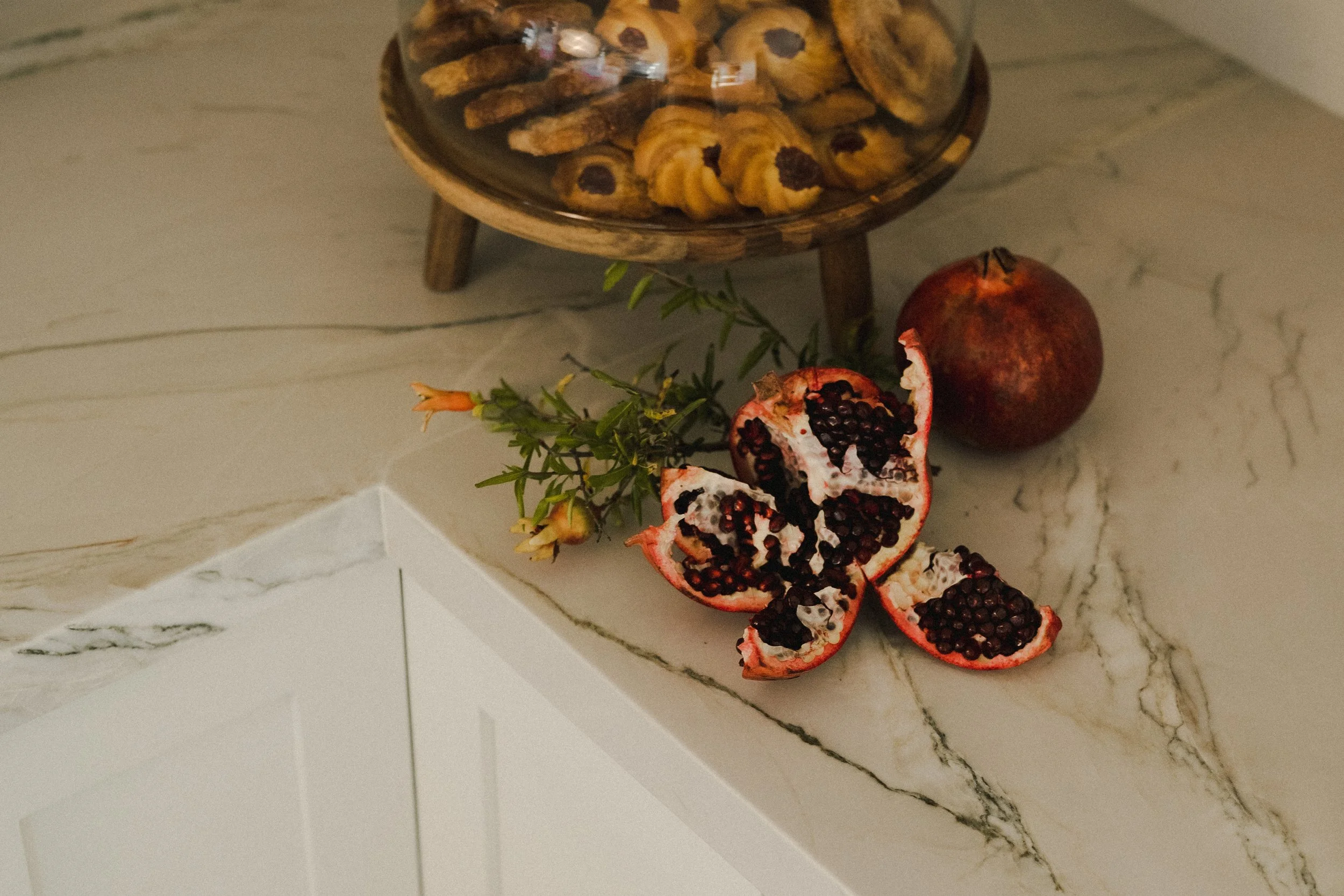 A marble countertop with a sprig of greenery, a pomegranate cut open revealing seeds, another whole pomegranate, and a small wooden stand with a glass dome cover containing assorted cookies.
