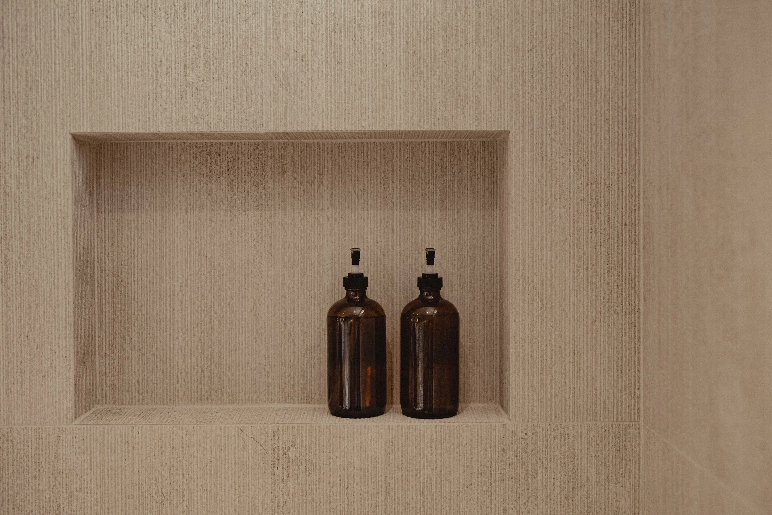 Two dark amber bottles with black caps on a built-in wall shelf in a beige tiled shower.