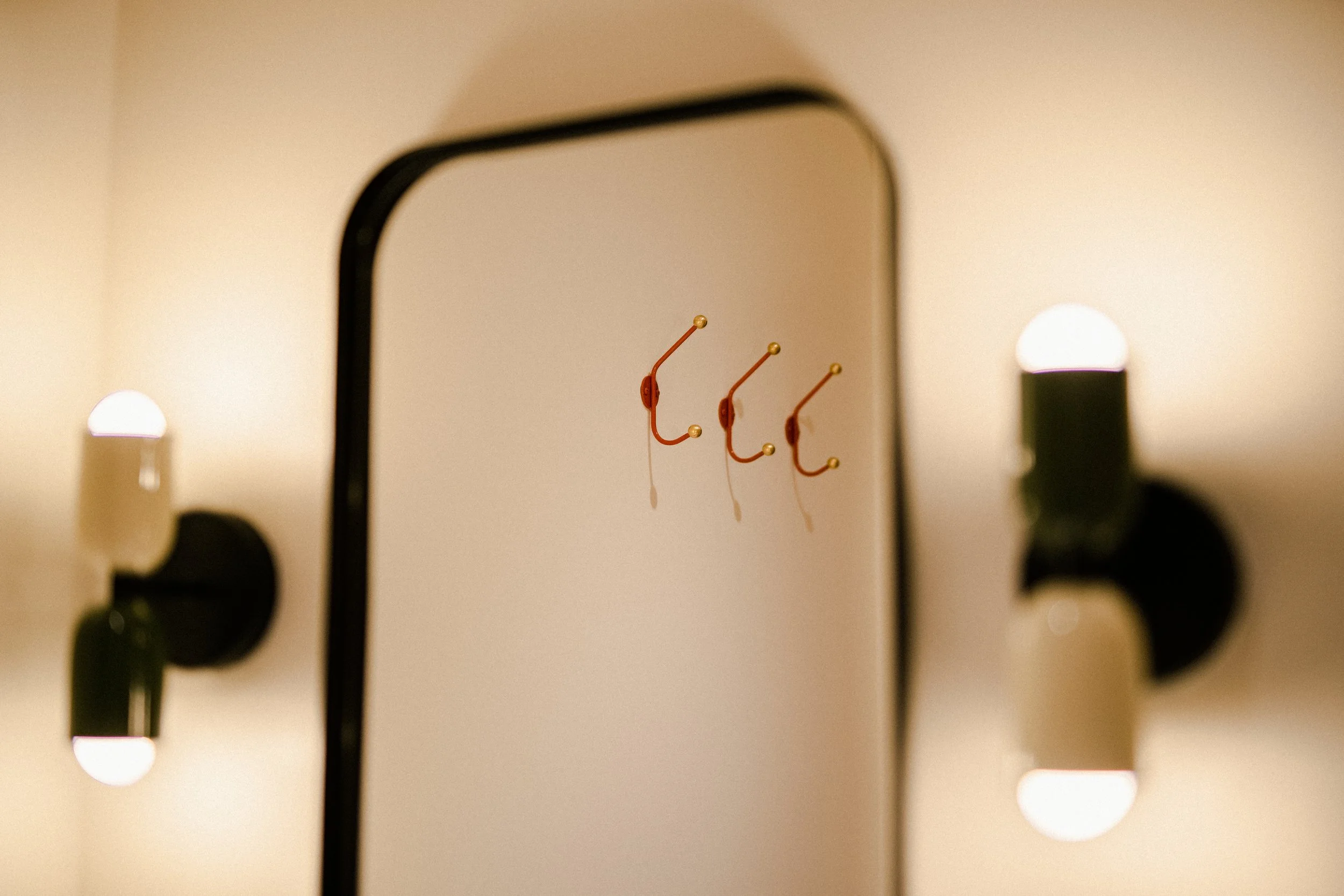 A wall-mounted mirror with a reflection of three orange hooks with gold tips in a room with warm lighting.