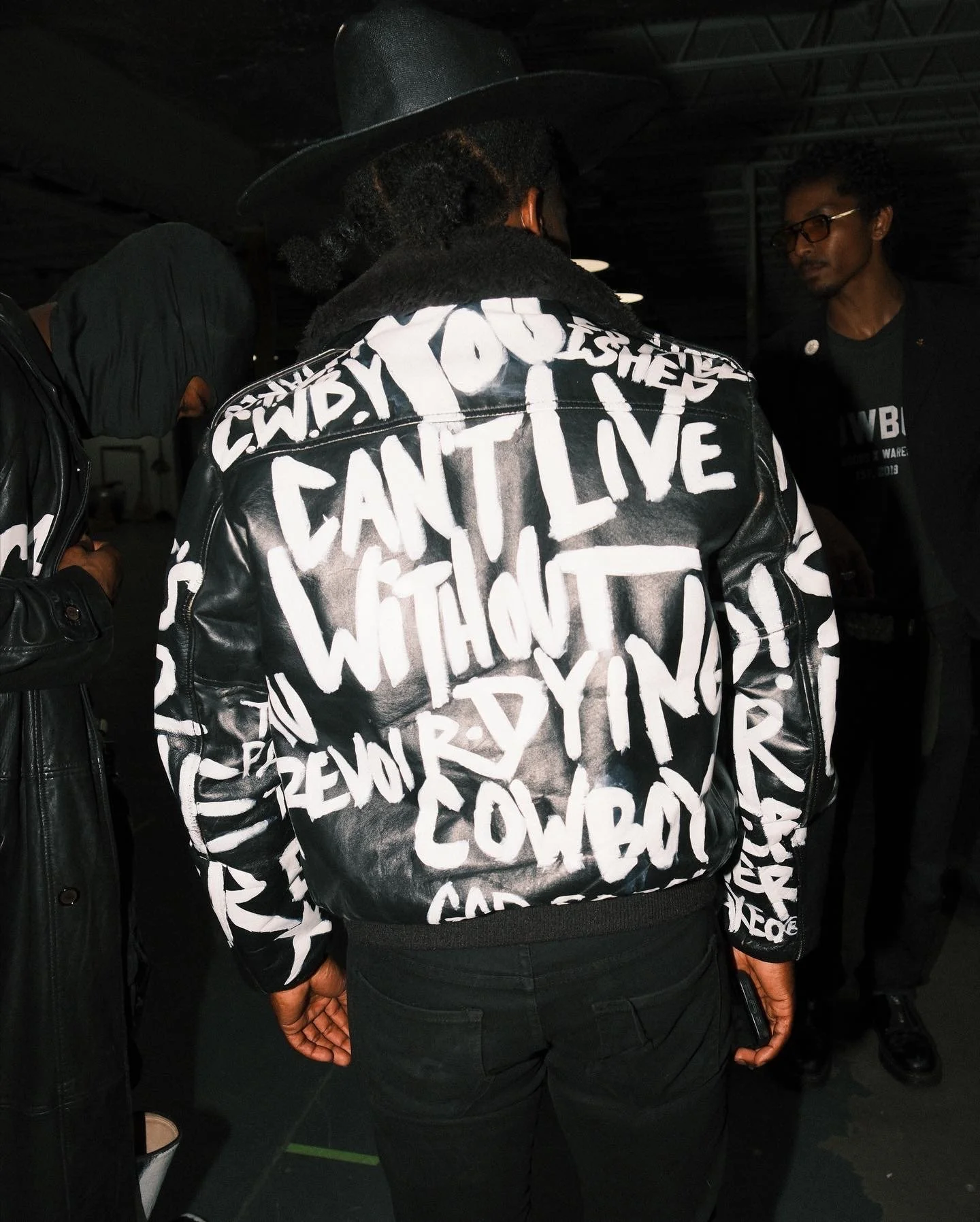 Person wearing a black leather jacket with white graffiti-style text in an indoor setting, with another person in the background.