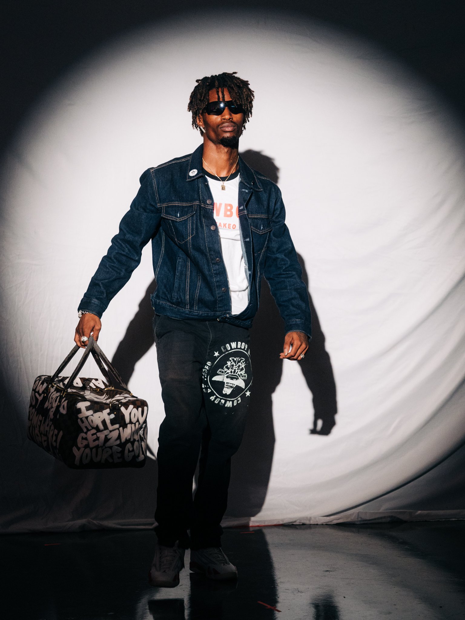 A man with dreadlocks and sunglasses walking, carrying a patterned duffel bag, wearing a denim jacket, black sweatpants, a white graphic t-shirt, and sneakers, against a plain backdrop.