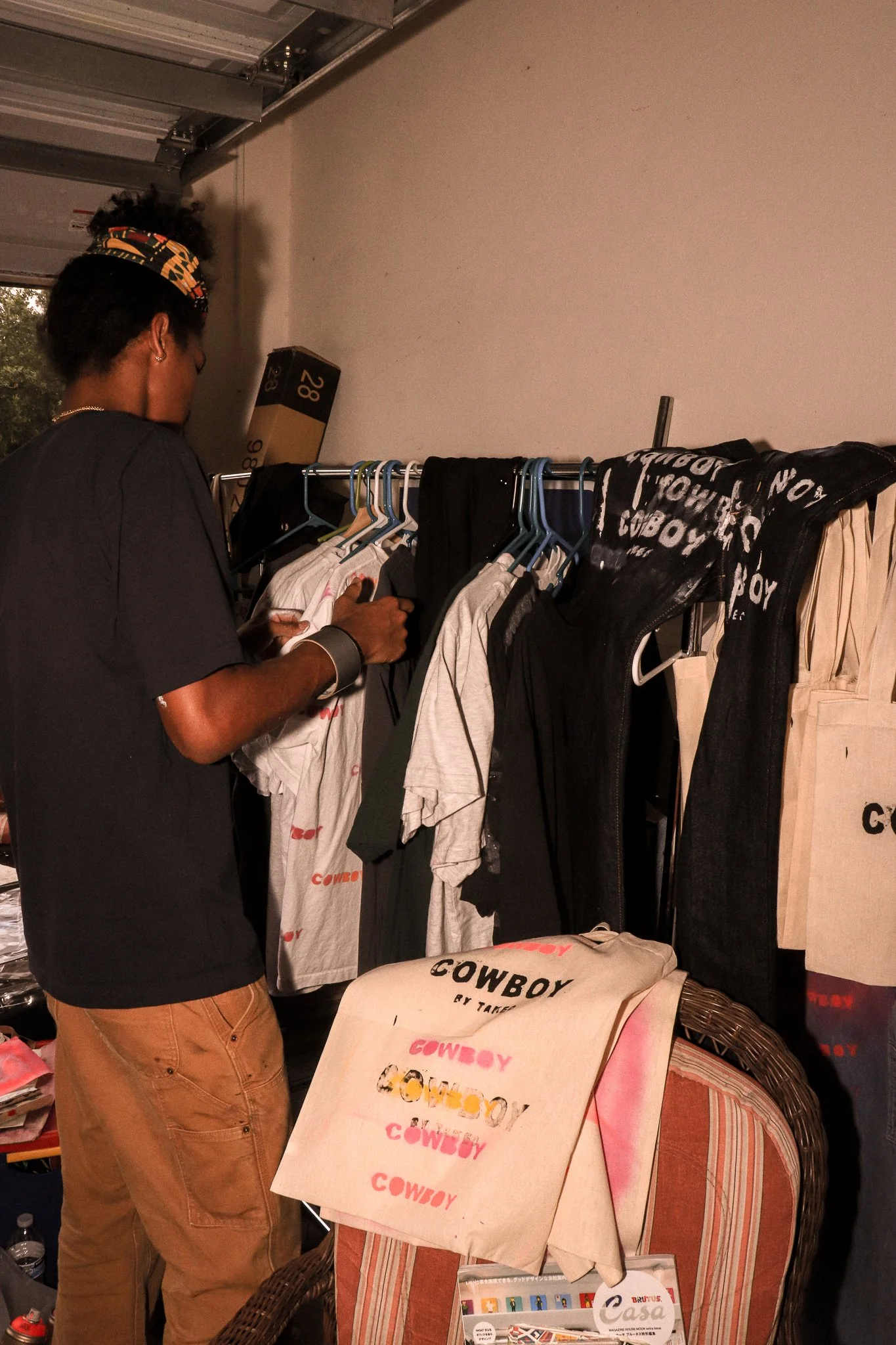 A person browsing through clothing items on a rack in a store or at a market, with T-shirts and bags featuring the word 'COWBOY' displayed.