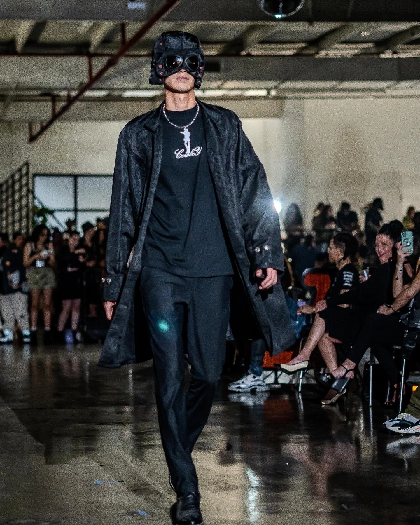 Male model walking on runway at fashion show, wearing black sunglasses, black coat, black shirt with 'Cowboy' logo, and black pants, audience seated on sides.