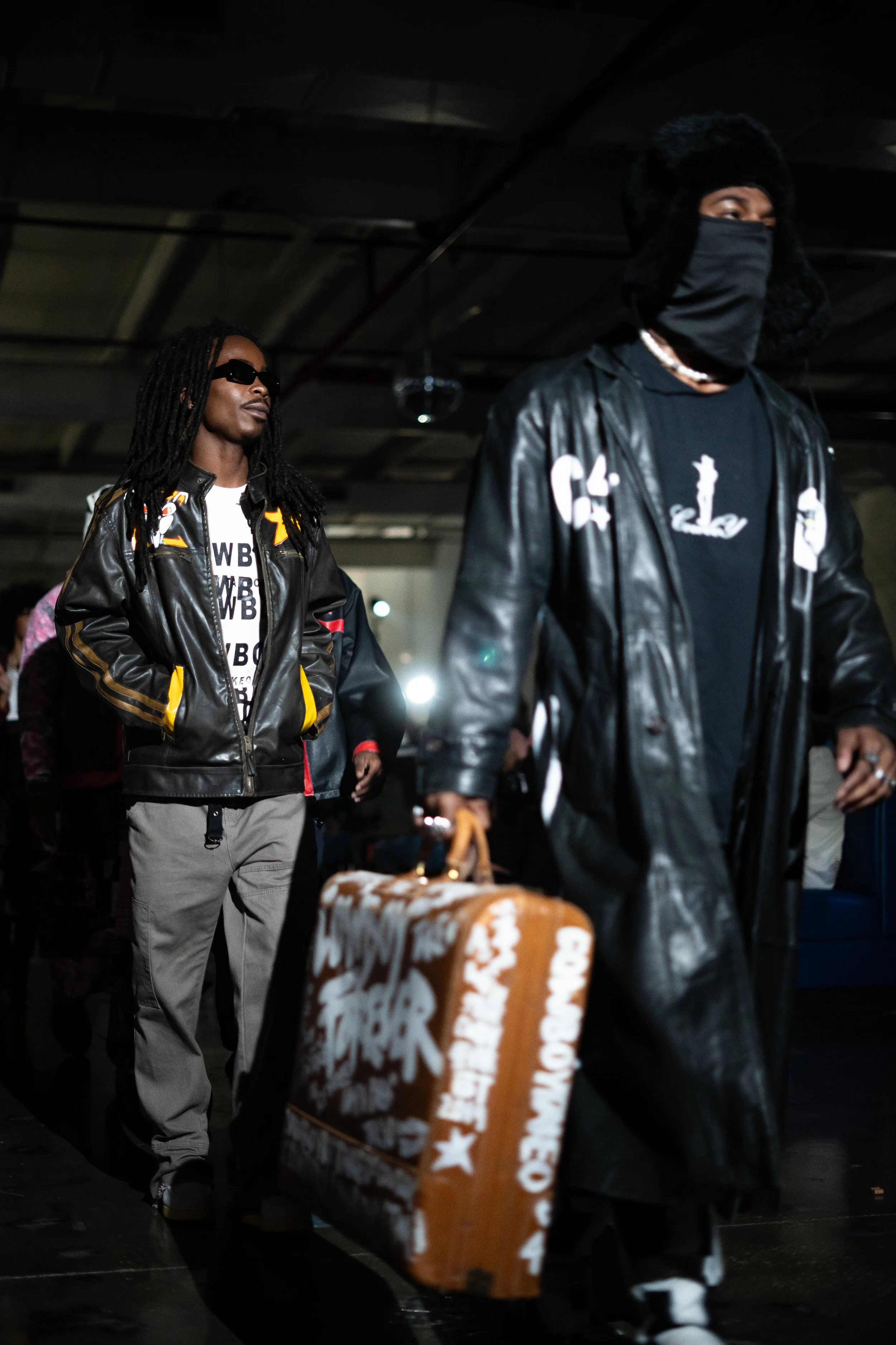 Two men walking in an indoor space, with one carrying a large brown box with white text and graphics, both wearing black leather jackets and face coverings.