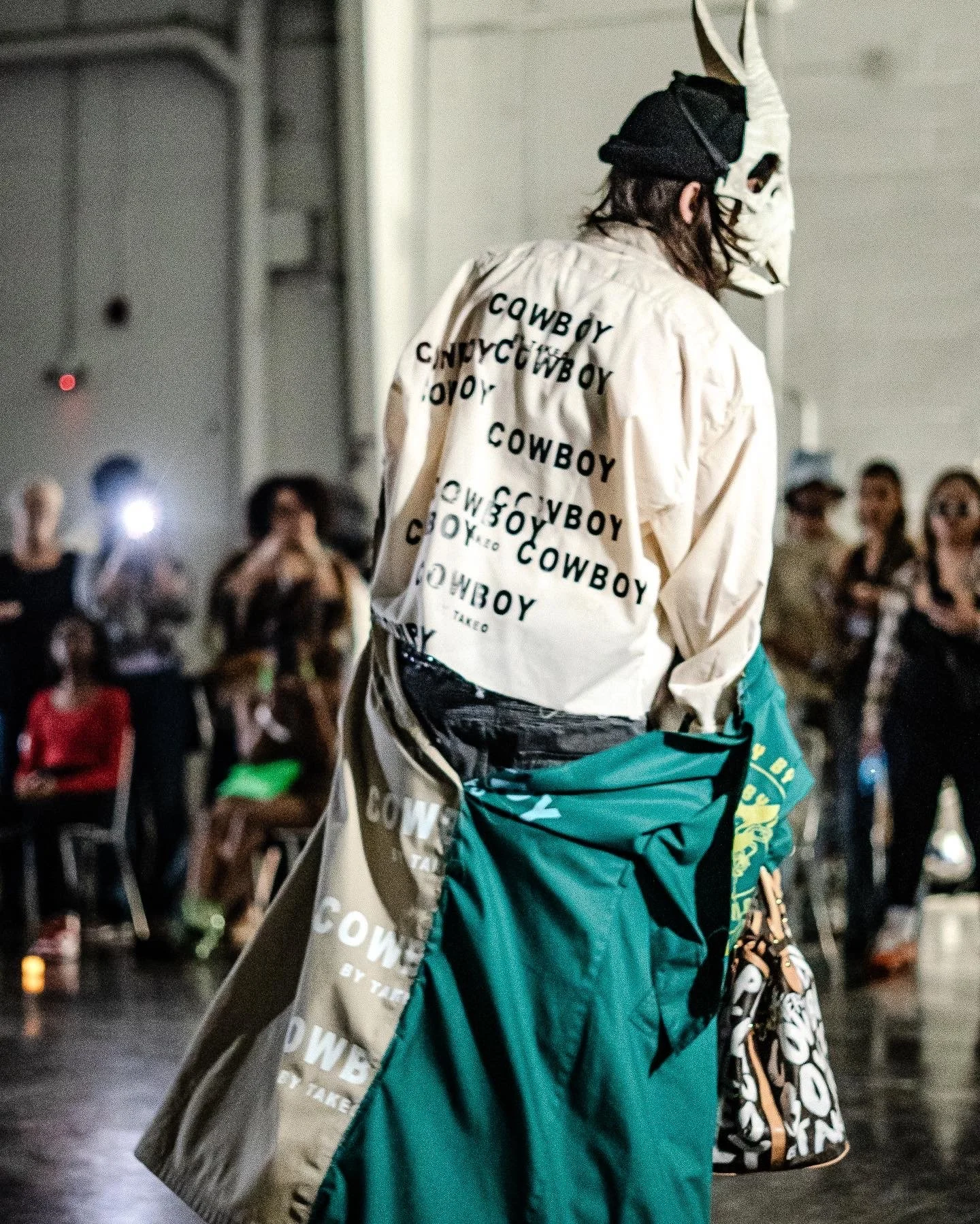 Person wearing a white jacket with the word 'COWBOY' repeated multiple times, a black hat, a mask resembling a skull, and carrying a large teal bag, standing in front of an audience in an indoor setting.