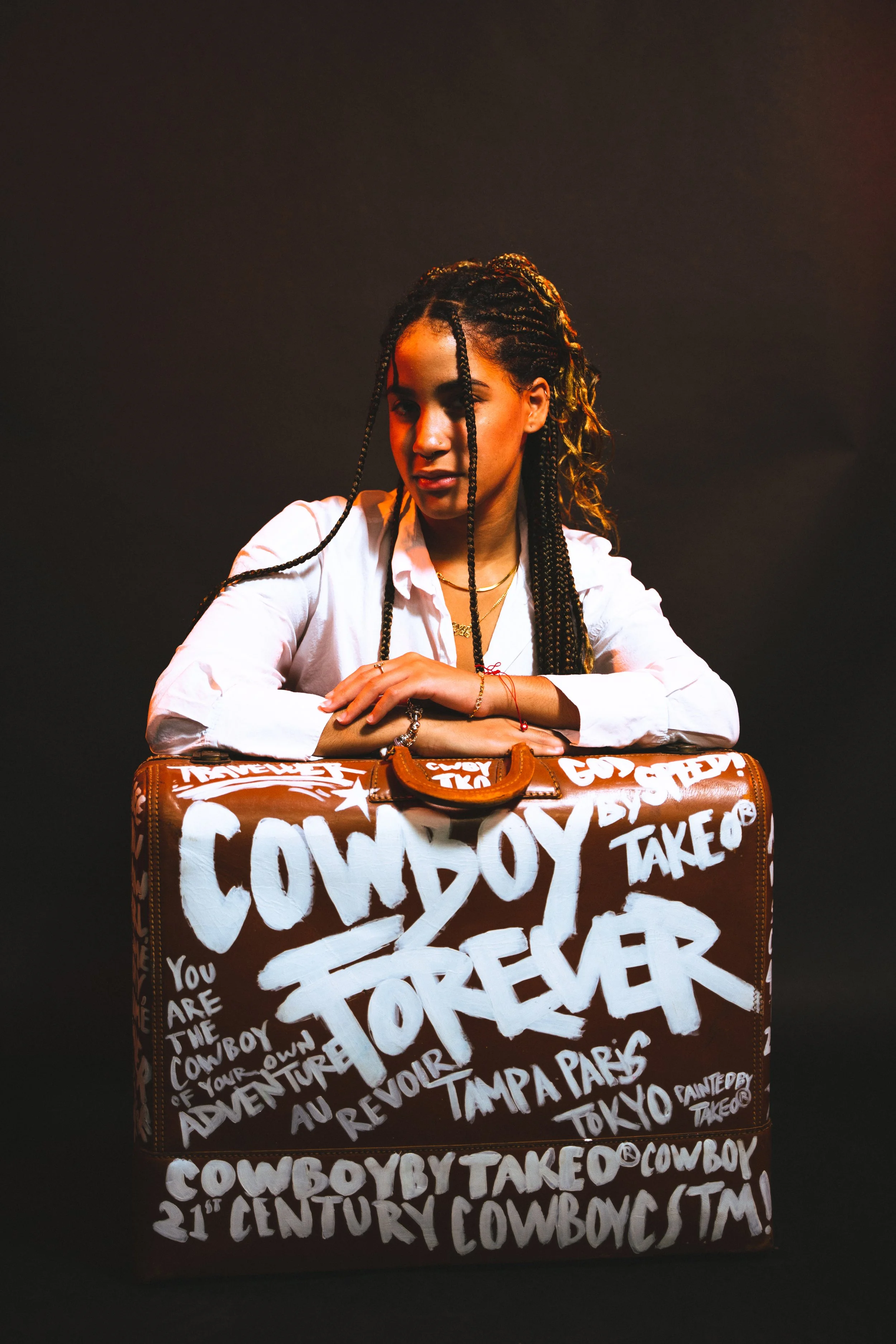 A woman with braided hair and wearing a white shirt, leaning on a brown suitcase with white graffiti-style text and phrases about cowboys and the wild west, against a dark background.