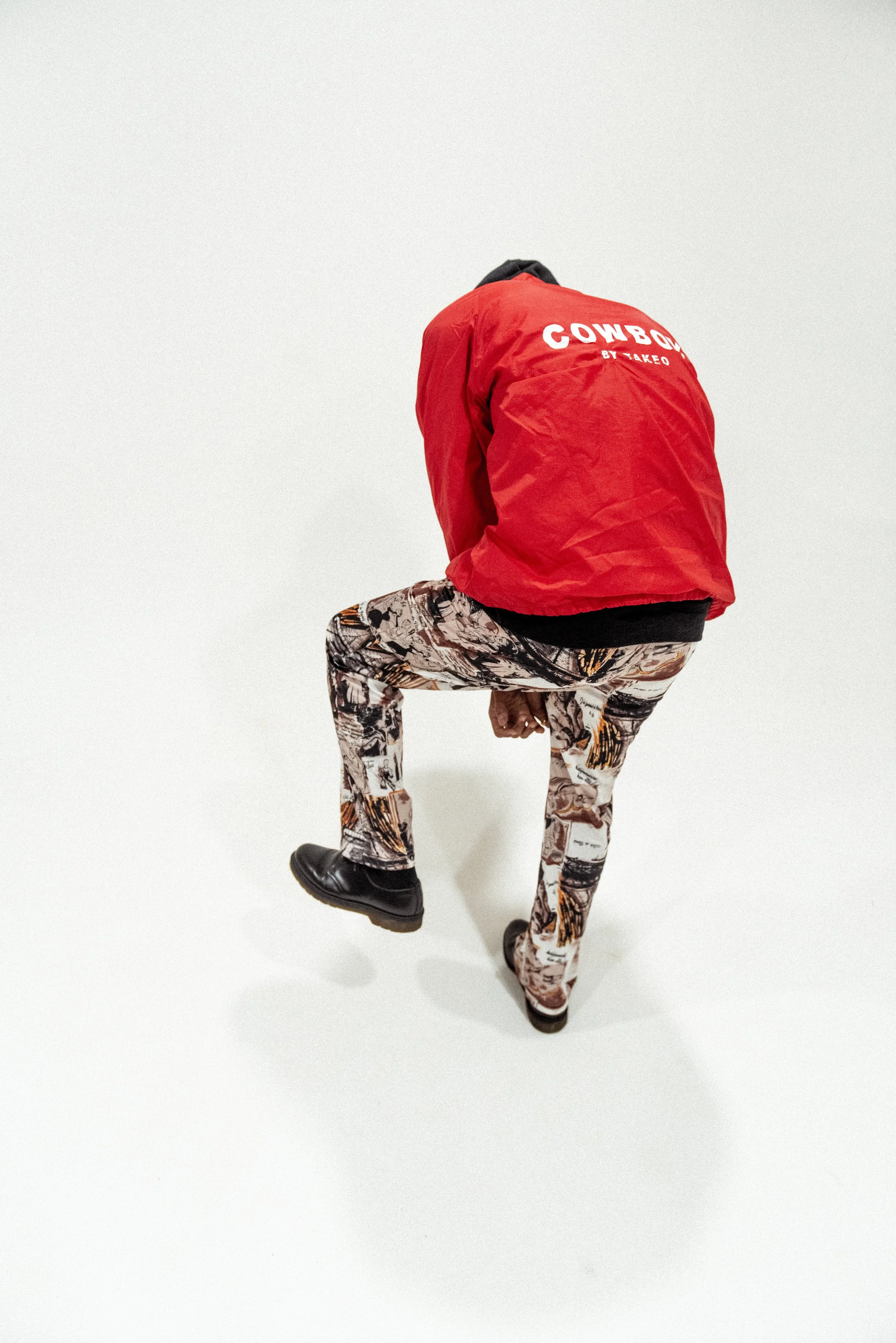 Person dressed in a red jacket with "Cowboy" written on the back, patterned pants, and black shoes, standing on a white background.