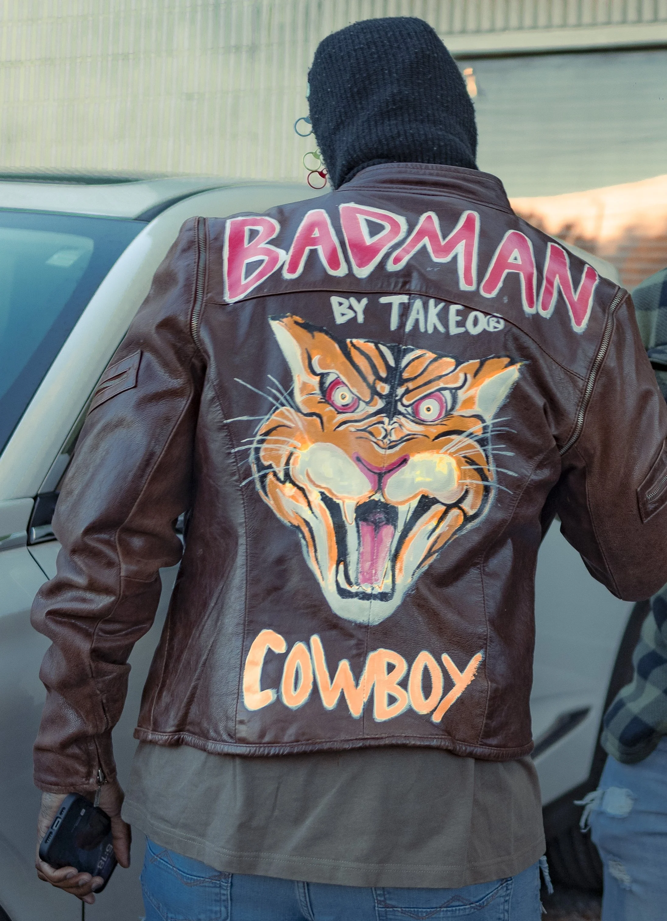 Back of a person wearing a brown leather jacket with colorful patches and text, standing near a white vehicle.