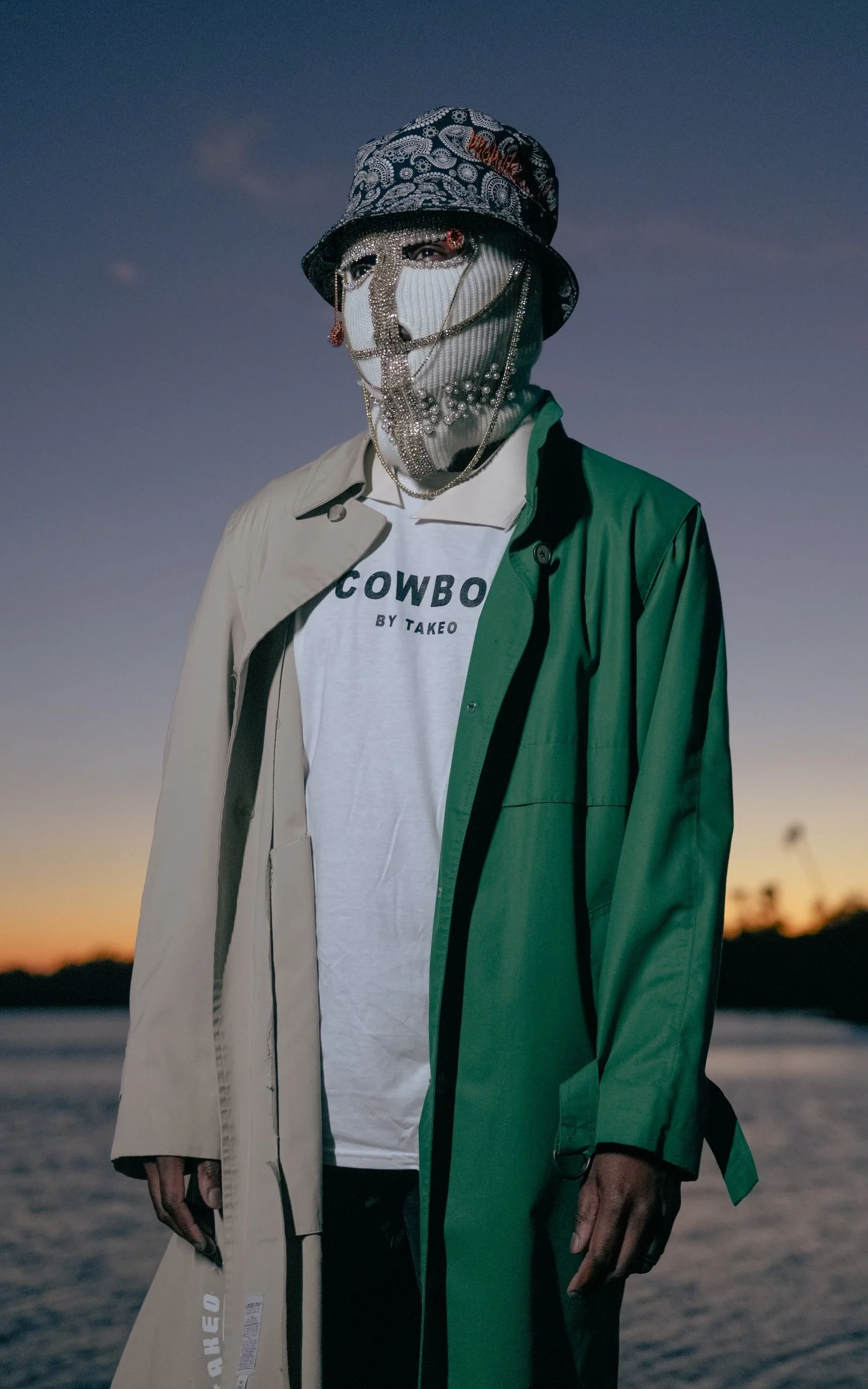 Person wearing a face mask, embellished with jewels, a patterned bucket hat, and layered necklaces, standing outdoors at sunset near a body of water, dressed in a T-shirt and two-tone trench coat.